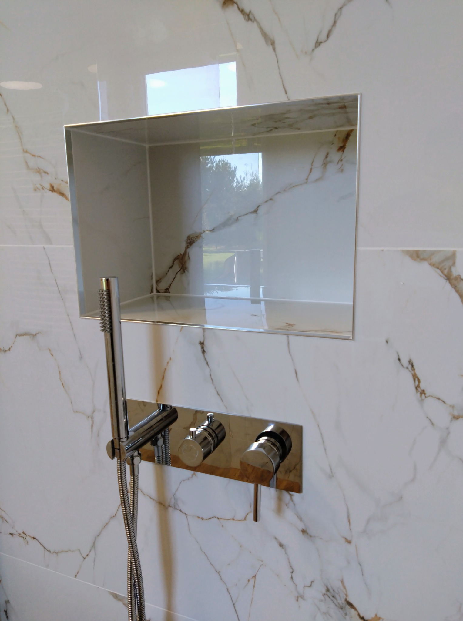 Biała ściana łazienki z marmurowym wzorem, wnęka z lustrzanym wykończeniem, bateria prysznicowa z chromowanymi elementami. Nowoczesny design.