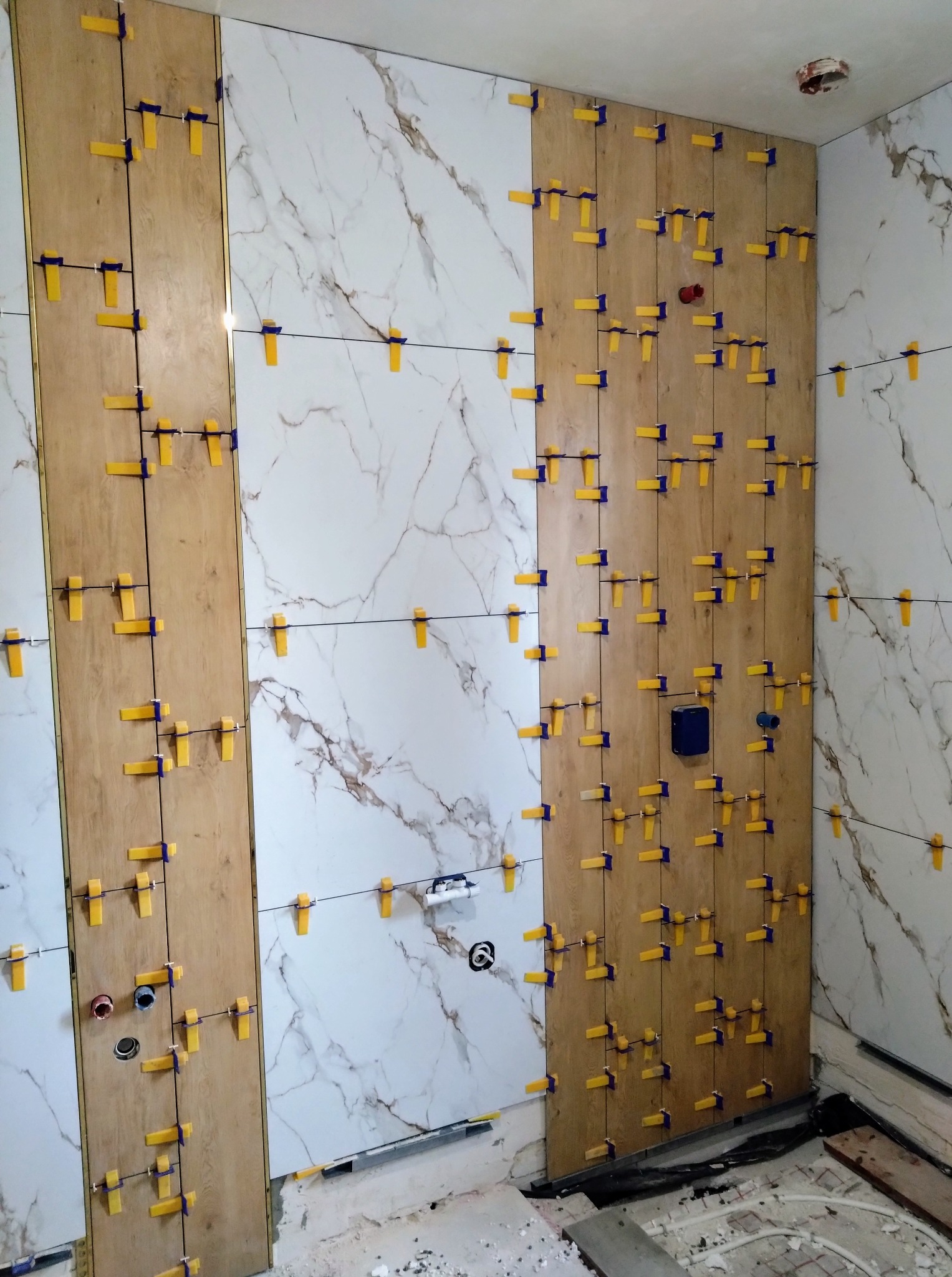 Układanie płytek imitujących drewno i marmur na ścianie z użyciem klipsów poziomujących. Widoczne otwory na instalacje i fragment podłogi z ogrzewaniem.