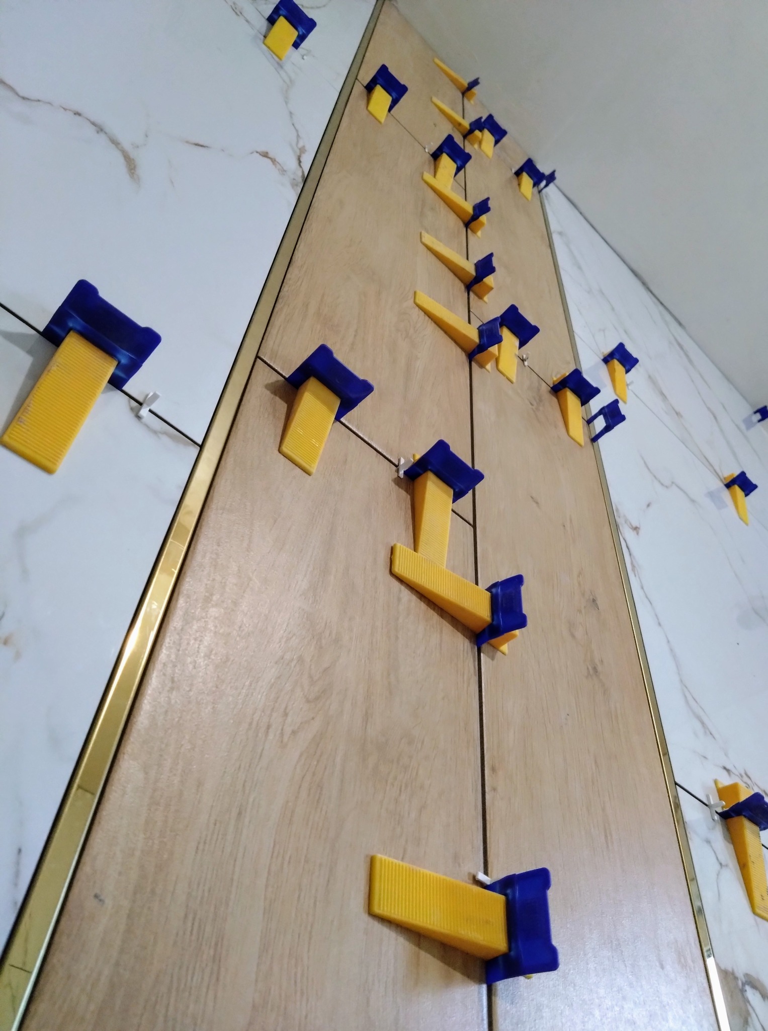 Układanie płytek imitujących drewno i marmur z użyciem systemu poziomowania. Widoczne żółto-niebieskie klipsy i złota listwa wykończeniowa.