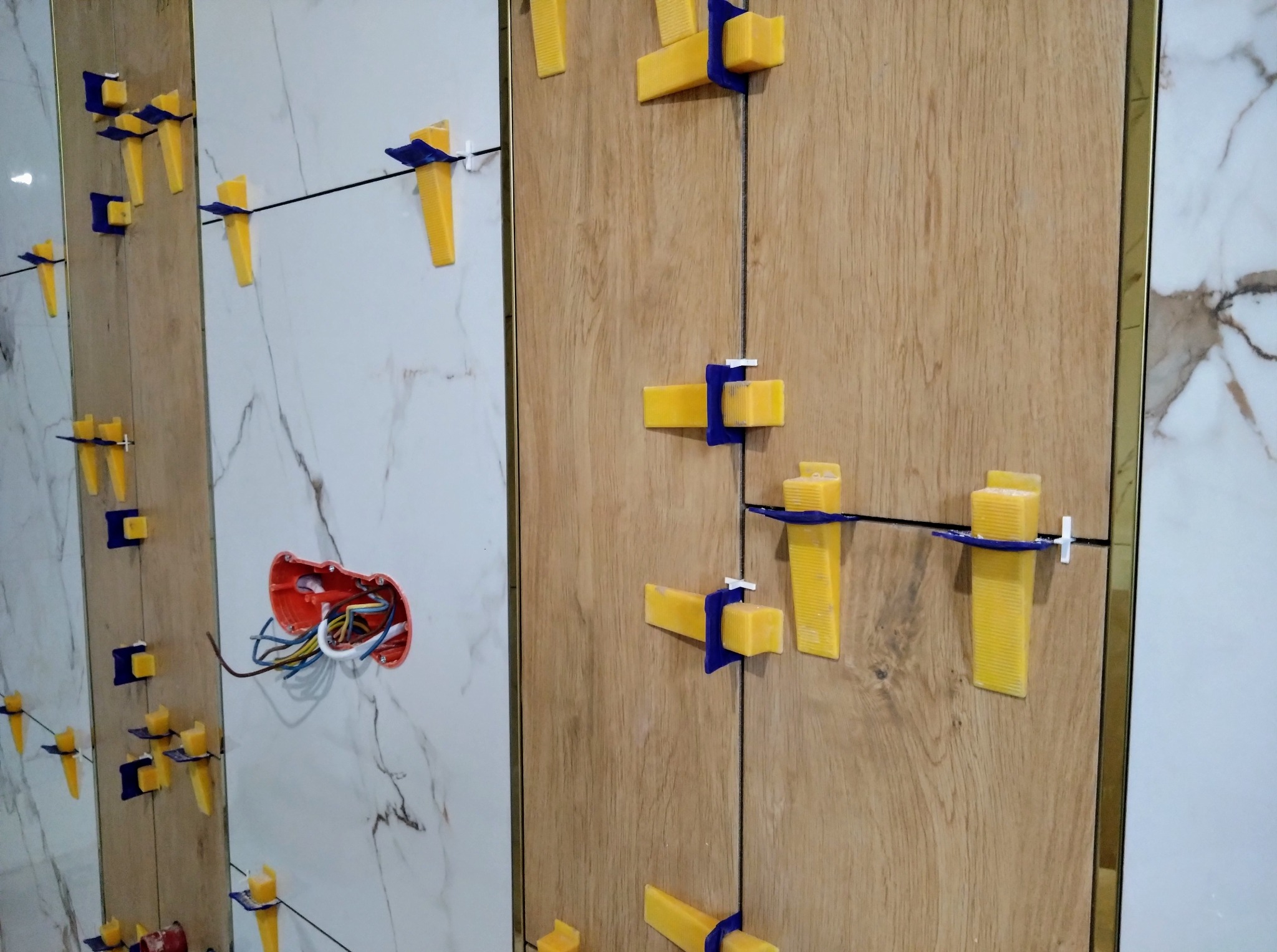 Układanie płytek imitujących drewno i marmur z użyciem systemu poziomowania. Widoczne kable w puszce elektrycznej. Detal z bliska, oświetlenie naturalne.