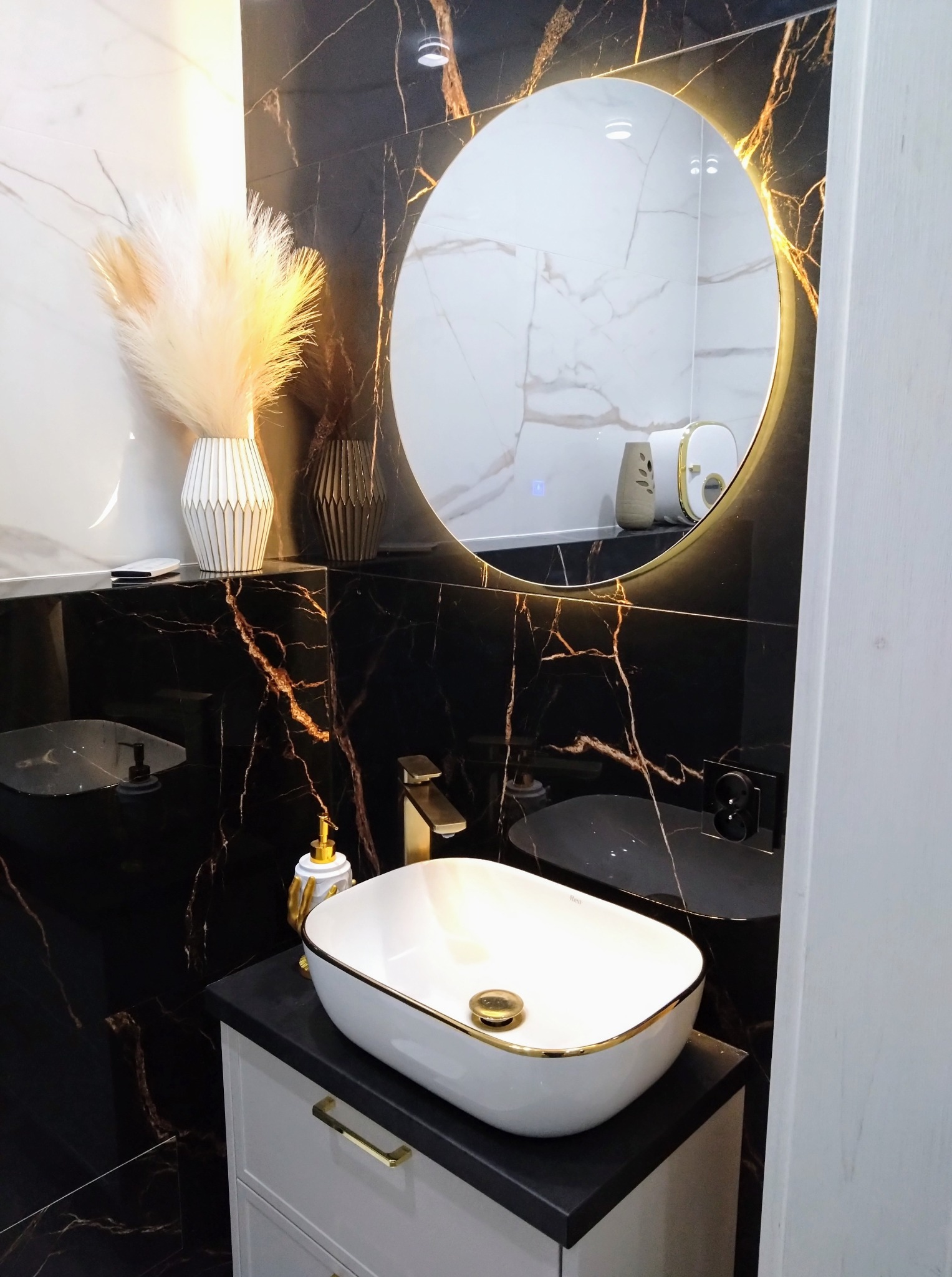 Elegancka łazienka z czarnymi płytkami imitującymi marmur, owalną umywalką nablatową z złotymi akcentami i okrągłym lustrem z podświetleniem LED.