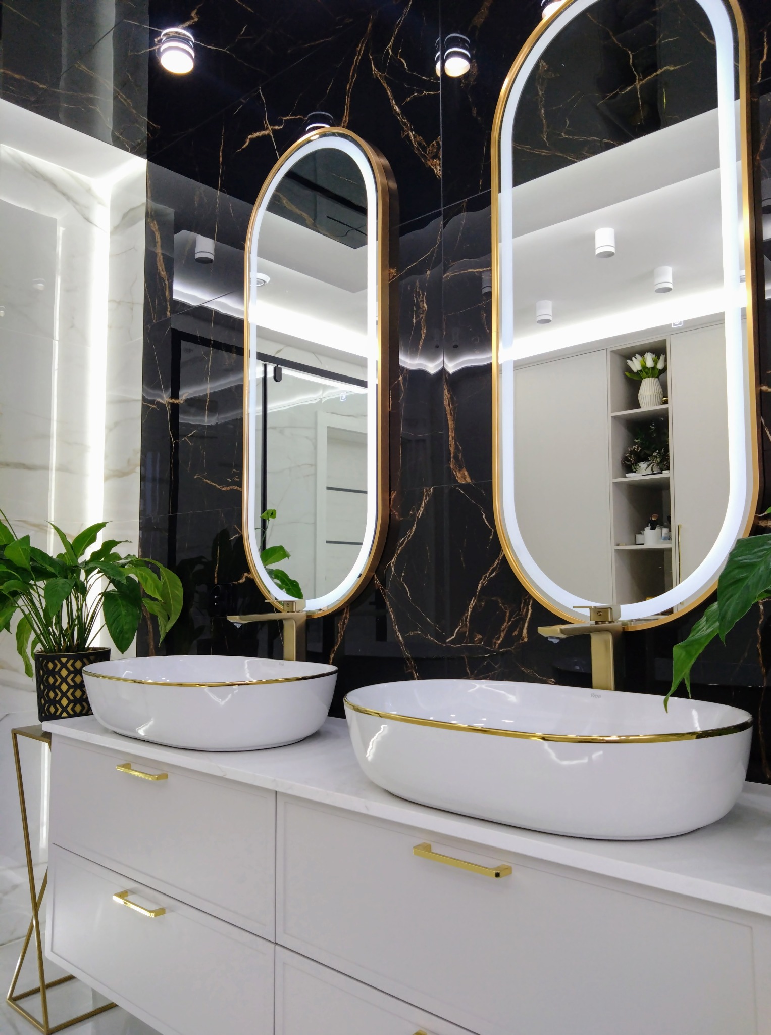 Elegancka łazienka z dwoma owalnymi umywalkami w złotym wykończeniu, podświetlanymi lustrami i czarnymi płytkami imitującymi marmur.