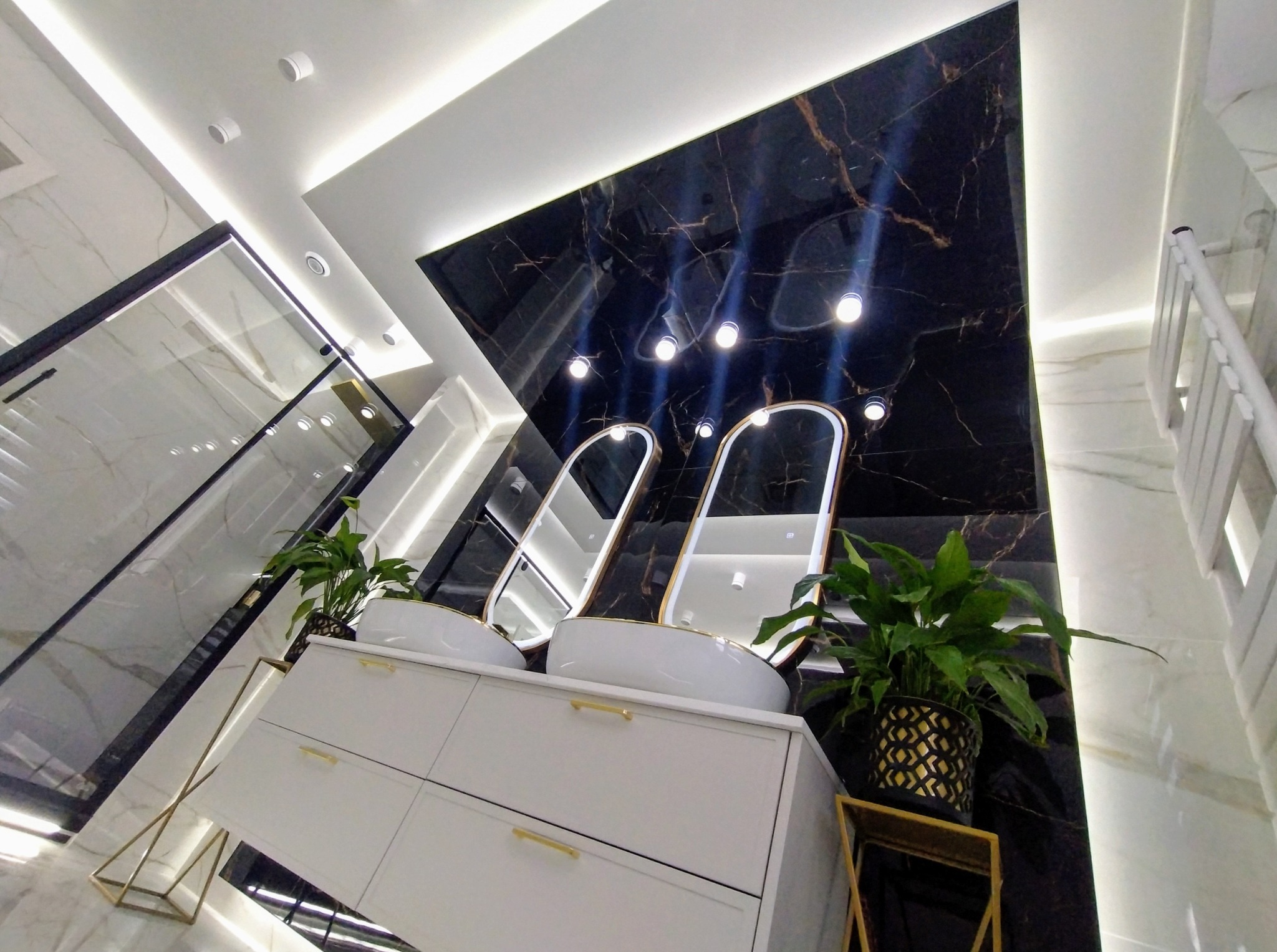 Nowoczesna łazienka z czarnym marmurem, białymi szafkami i umywalkami nablatowymi. Oświetlenie punktowe i rośliny doniczkowe dodają elegancji.