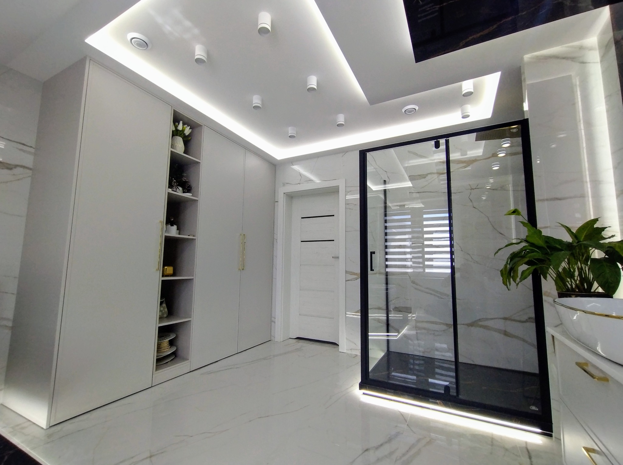 Nowoczesna łazienka z kabiną prysznicową typu walk-in, marmurową podłogą i zabudową meblową w odcieniach bieli. Oświetlenie sufitowe punktowe i liniowe LED.