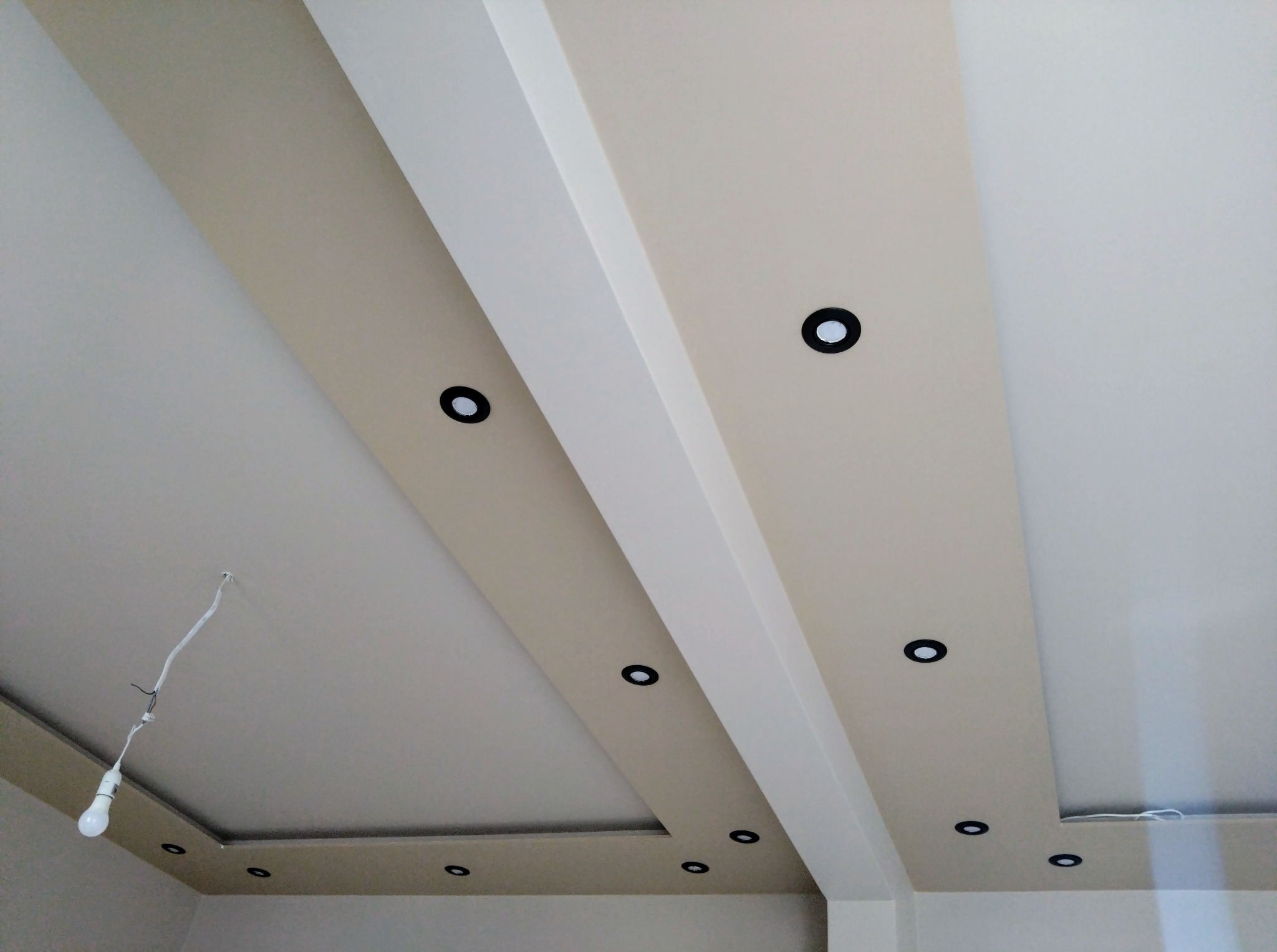 Wykończony sufit podwieszany z oświetleniem punktowym, widok pod kątem. Elementy dekoracyjne w odcieniach bieli i beżu. Wisząca żarówka na kablu w lewym dolnym rogu.