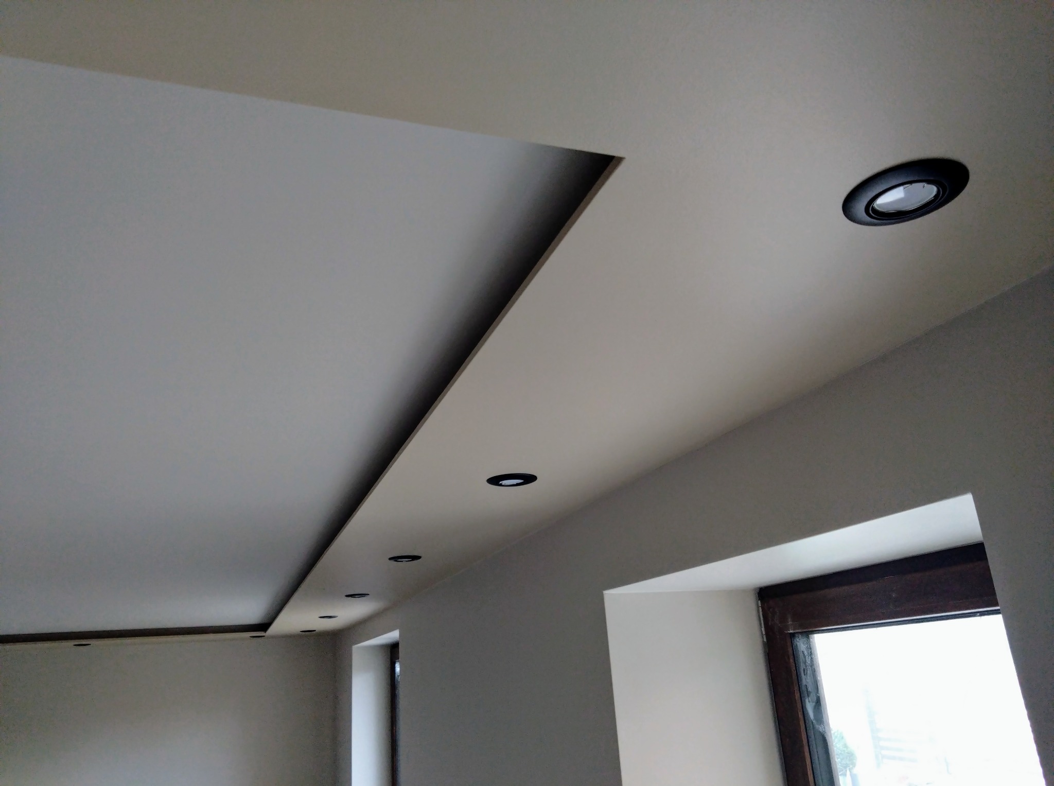 Nowoczesny sufit podwieszany z oświetleniem punktowym w jasnym wnętrzu. Widoczne okno z ciemną ramą i fragment ściany. Minimalistyczny design.