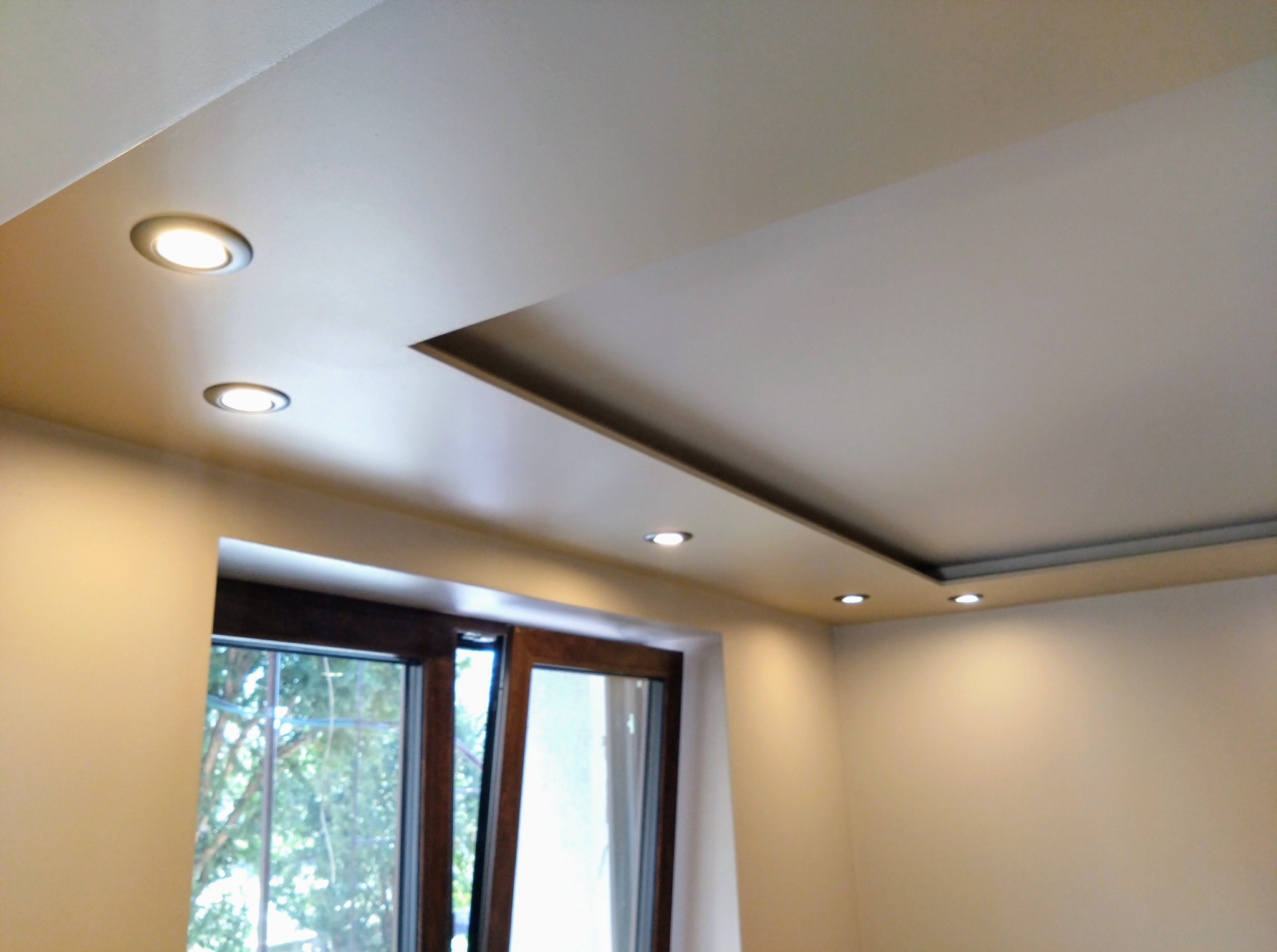 Podwieszany sufit z oświetleniem punktowym i wnęką nad oknem, świeżo po remoncie. Widok na fragment pokoju z oknem.