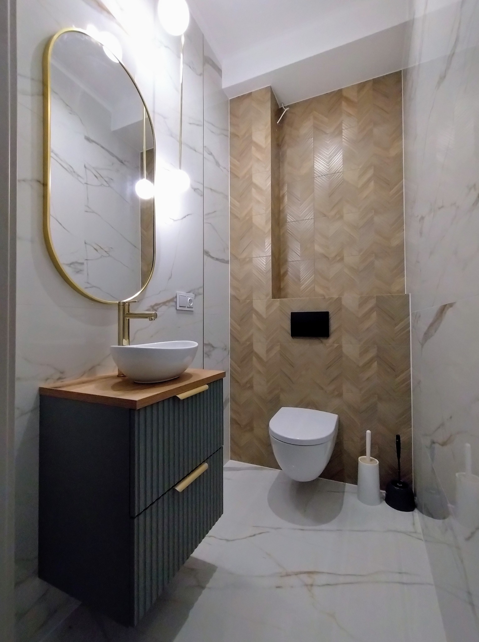 Nowoczesna łazienka po remoncie: marmurowe płytki, drewnopodobna ściana z wzorem jodełki, wisząca toaleta i umywalka nablatowa z lustrem w złotej ramie.