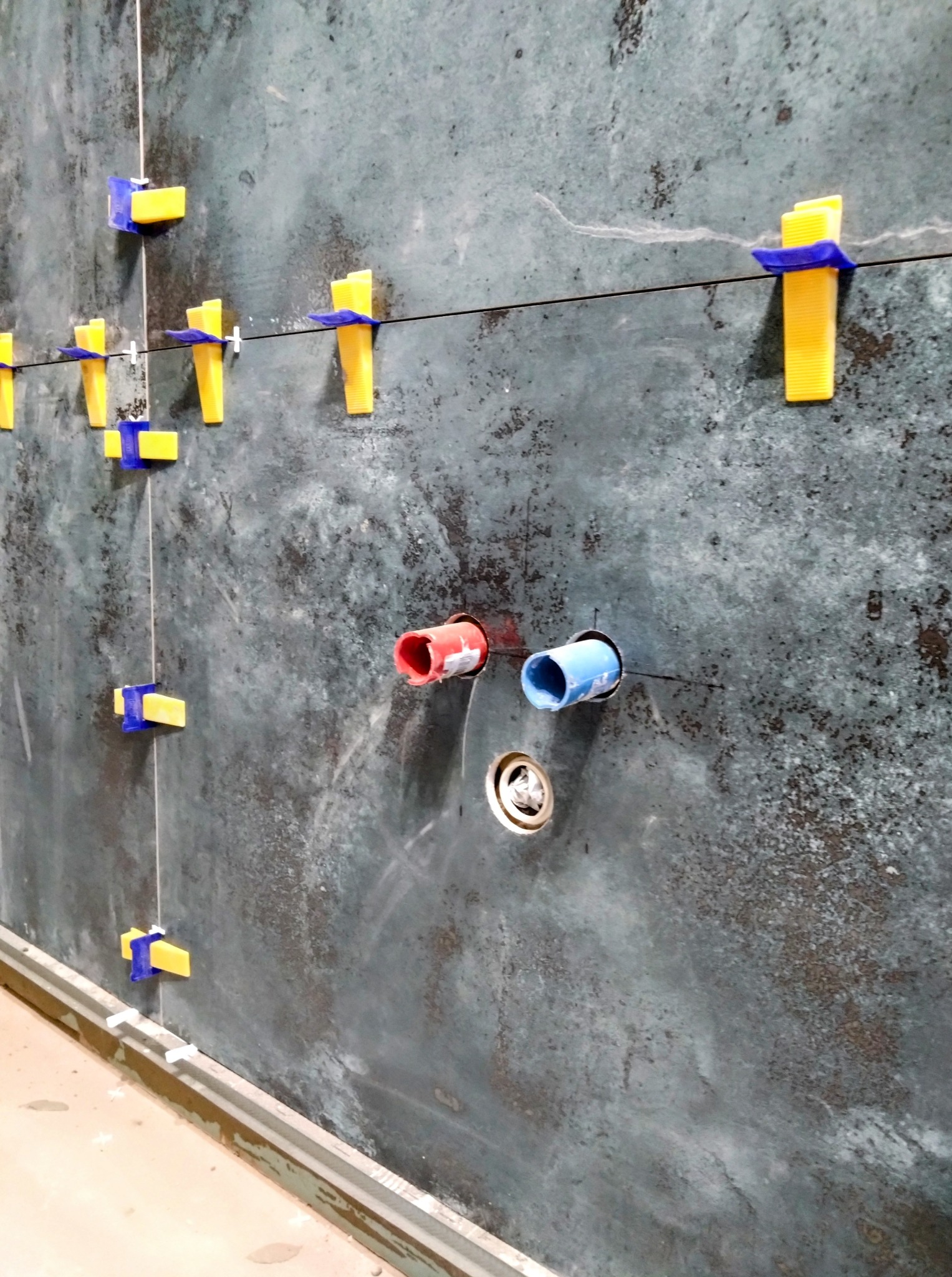 Ściana z szarych płytek, z żółto-niebieskimi klipsami poziomującymi, otwory na rury (czerwona i niebieska) i odpływ. Widok z bliska, etap prac glazurniczych.