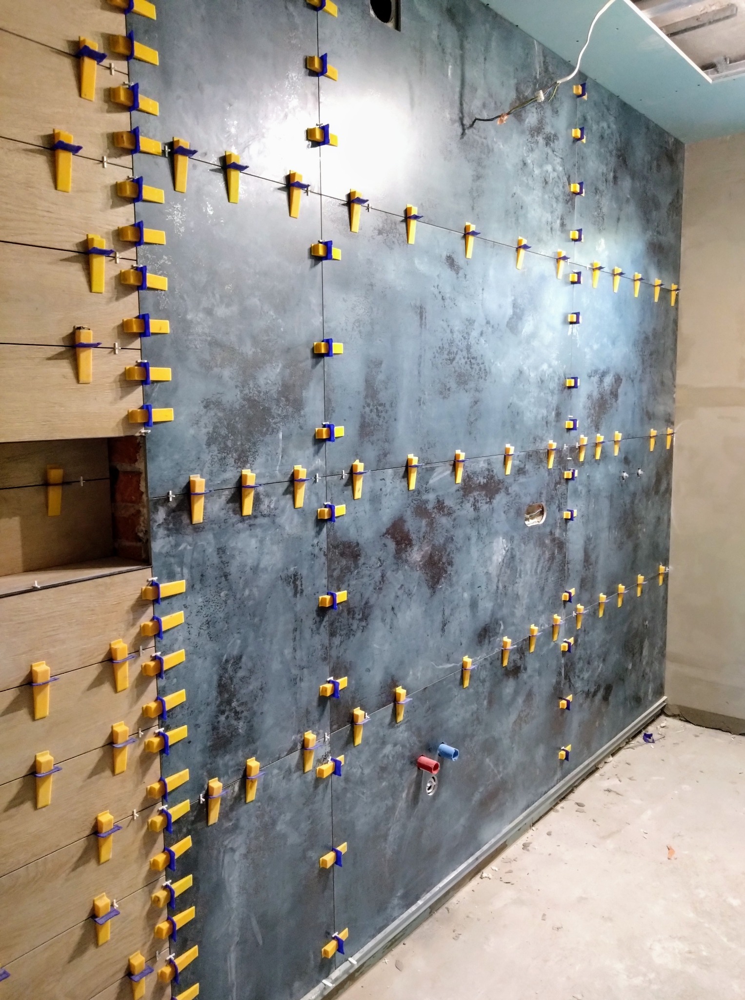 Układanie płytek na ścianie z użyciem systemu poziomowania; widoczne płytki drewnopodobne i szare, imitujące beton, z żółto-niebieskimi klipsami dystansowymi.