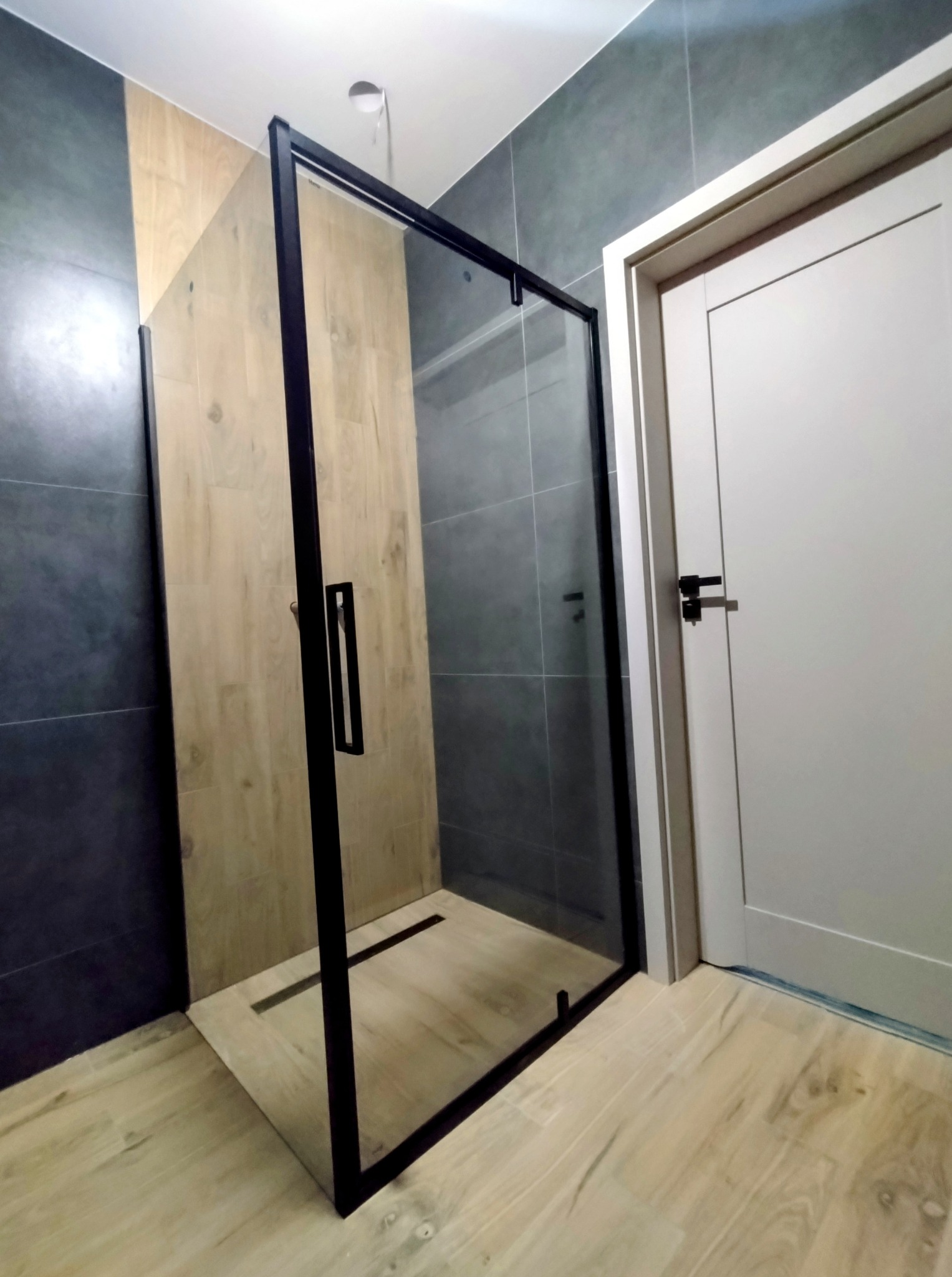Nowoczesna kabina prysznicowa z czarnymi profilami i odpływem liniowym, wykończona w Bełchatowie. Jasne płytki imitujące drewno, szare ściany i białe drzwi.