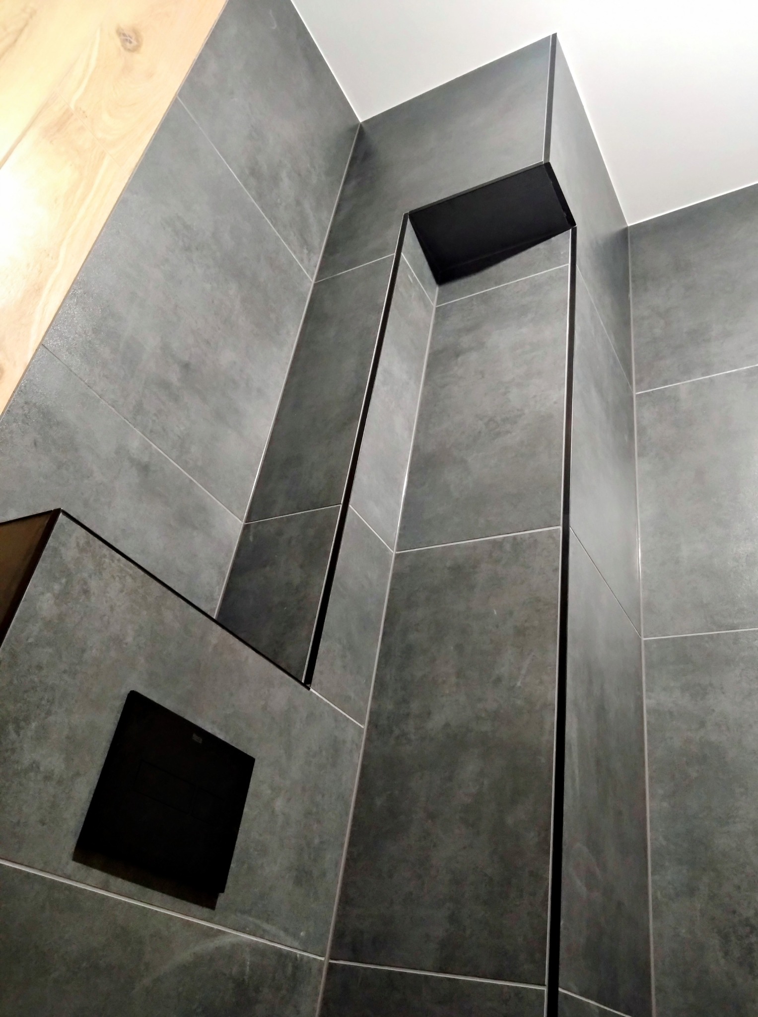 Szare, matowe płytki w łazience z czarnymi akcentami, tworzące nowoczesny design. Widoczna wnęka i kratka wentylacyjna. Ujęcie z dołu, podkreślające geometrię.