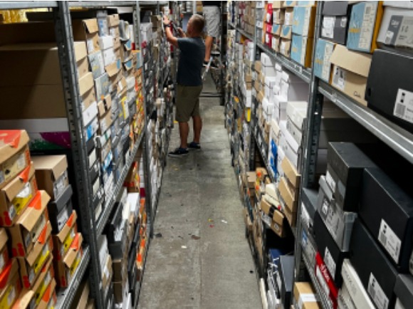 Wąski korytarz magazynowy z metalowymi regałami wypełnionymi kartonami, pracownik sięga po pudełko na górnej półce.