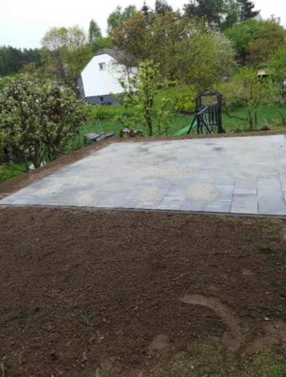 Przygotowany teren pod przyszłą nawierzchnię z kostki, widoczna wyrównana powierzchnia ziemi i częściowo ułożona kostka brzegowa, w tle dom z ogrodem.
