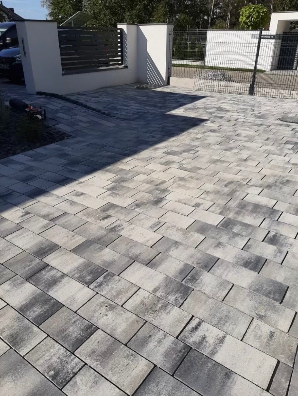 Ułożona szara kostka brukowa na podjeździe z widocznym fragmentem ogrodzenia panelowego z bramą w tle.