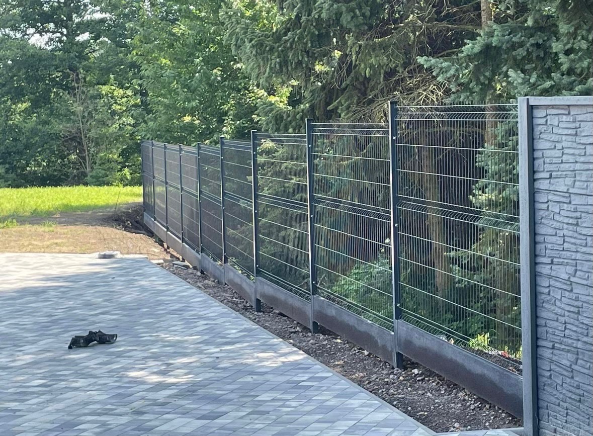 Nowoczesne ogrodzenie panelowe w kolorze grafitowym, z podmurówką na tle zieleni drzew, obok ułożona szara kostka brukowa.