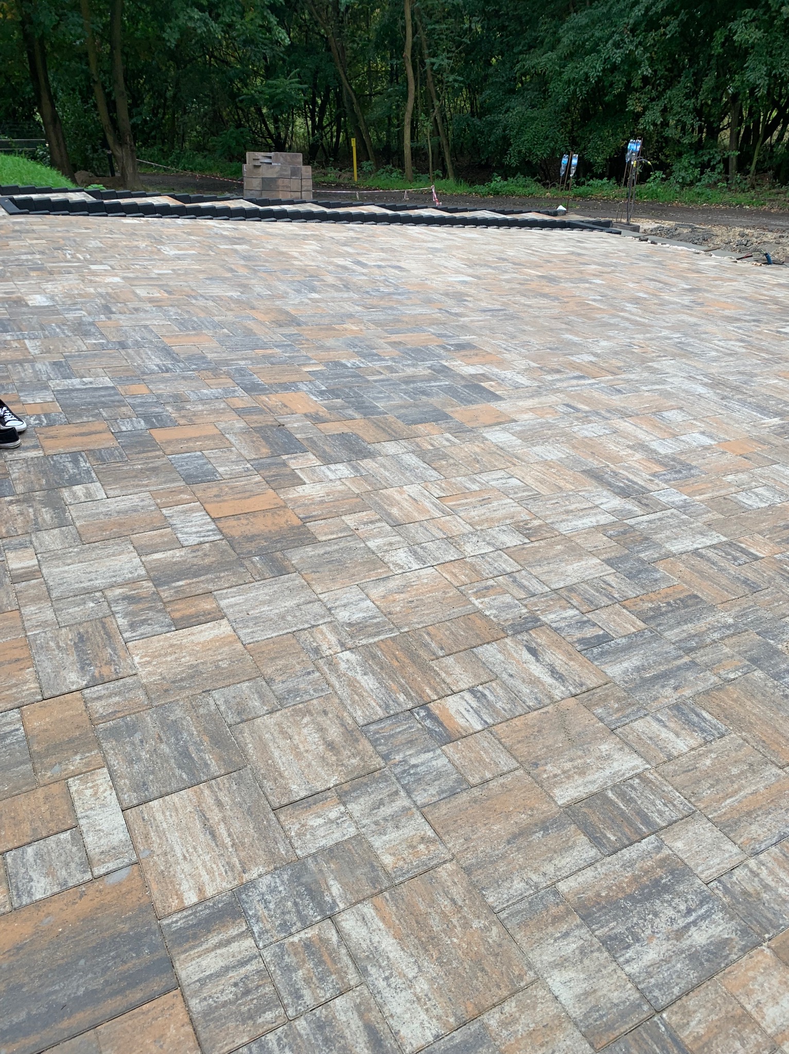 Ułożona kostka brukowa w odcieniach szarości i brązu, widok z góry, z elementami przygotowania podłoża i zapasem kostki w tle, na tle zieleni.