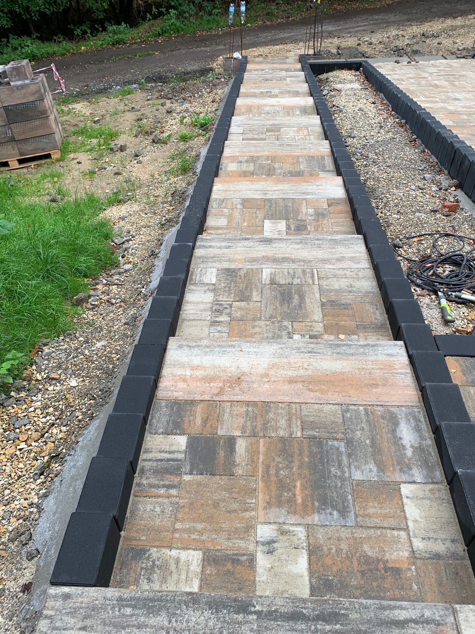 Ułożone schody z kostki brukowej w odcieniach szarości i beżu, obramowane czarnym obrzeżem. Widoczne narzędzia i paleta z kostką w tle.