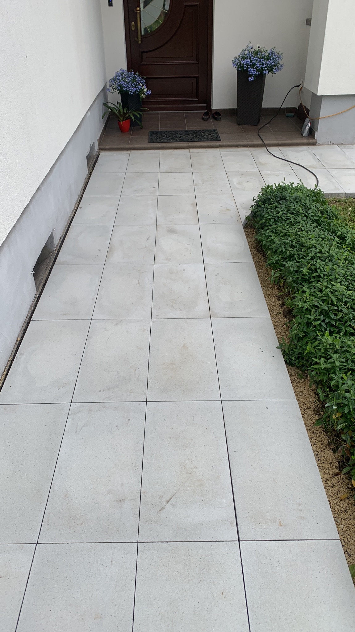Jasnoszara, kwadratowa kostka brukowa prowadząca do drzwi wejściowych, z roślinnością po prawej stronie. Widoczne donice z kwiatami przy drzwiach.