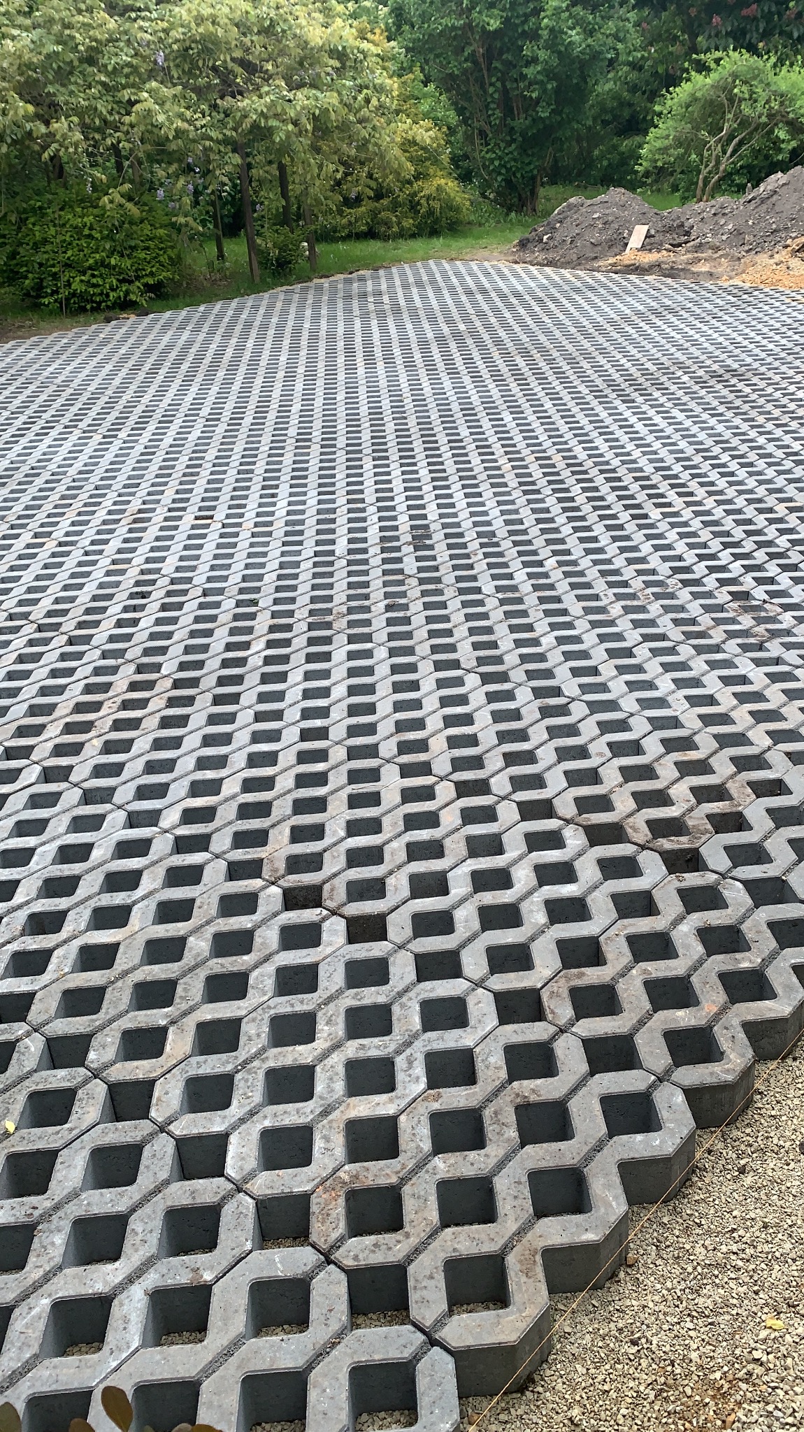 Szara, ażurowa nawierzchnia z betonowych płyt, tworząca wzór kraty na dużej powierzchni. W tle widoczna zieleń drzew i krzewów, oraz usypana ziemia.