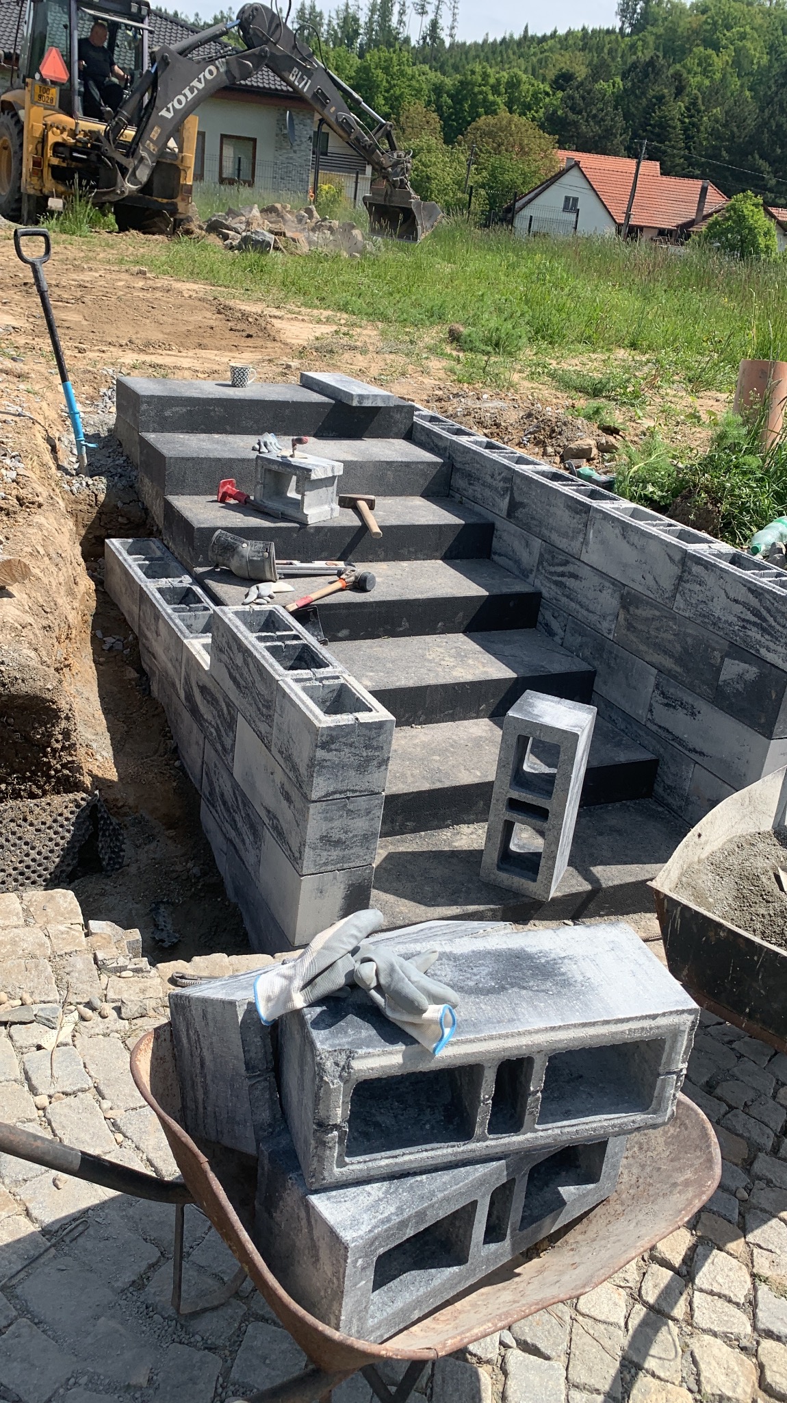 Budowa schodów zewnętrznych z betonowych bloczków, widoczne narzędzia i koparka w tle. Etap prac murarskich, częściowo ukończona konstrukcja.