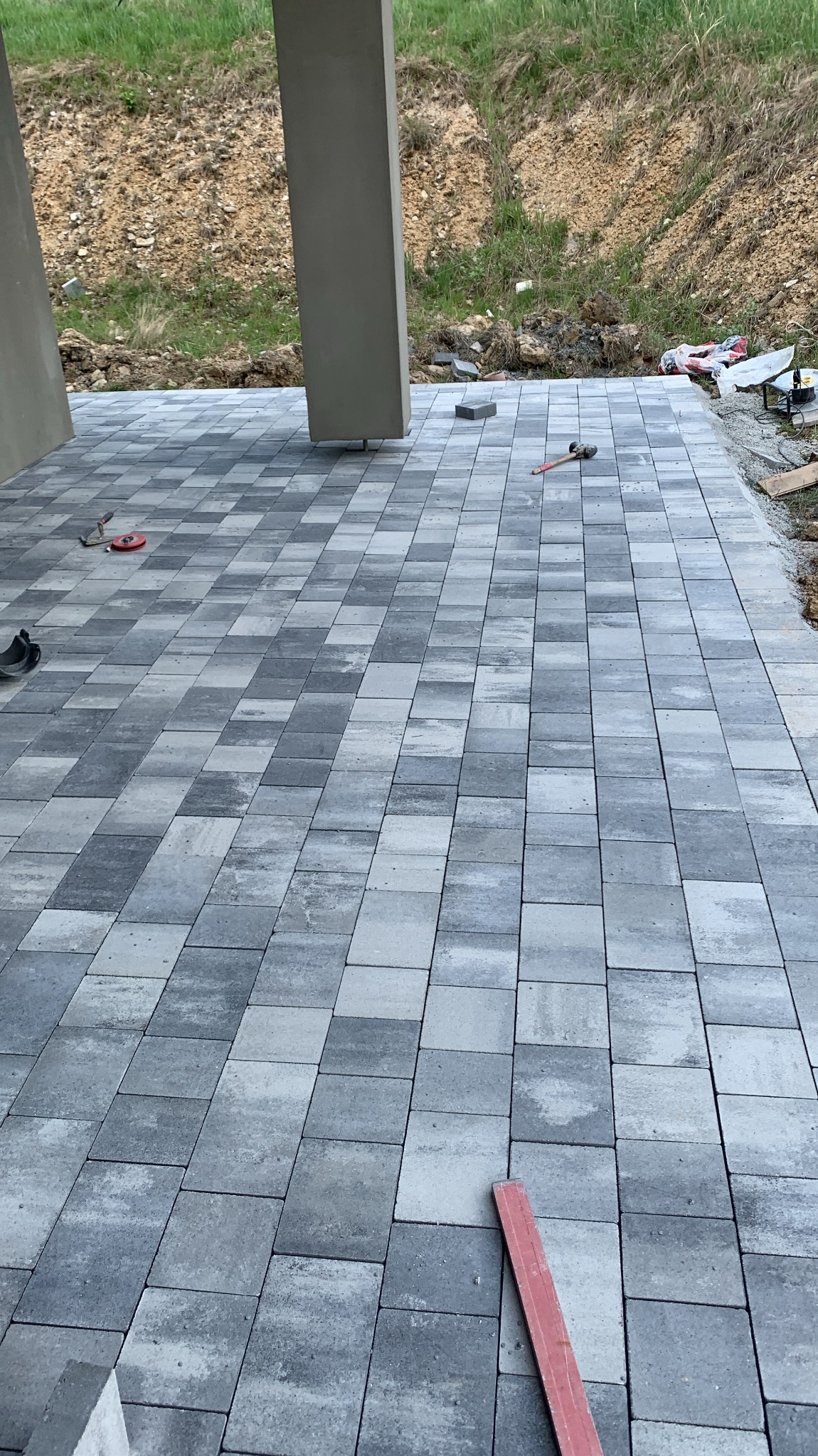 Ułożona, szara kostka brukowa na tarasie, widoczne narzędzia brukarskie i fragmenty konstrukcji budynku w tle. Regularny wzór kostki, świeżo po ułożeniu.