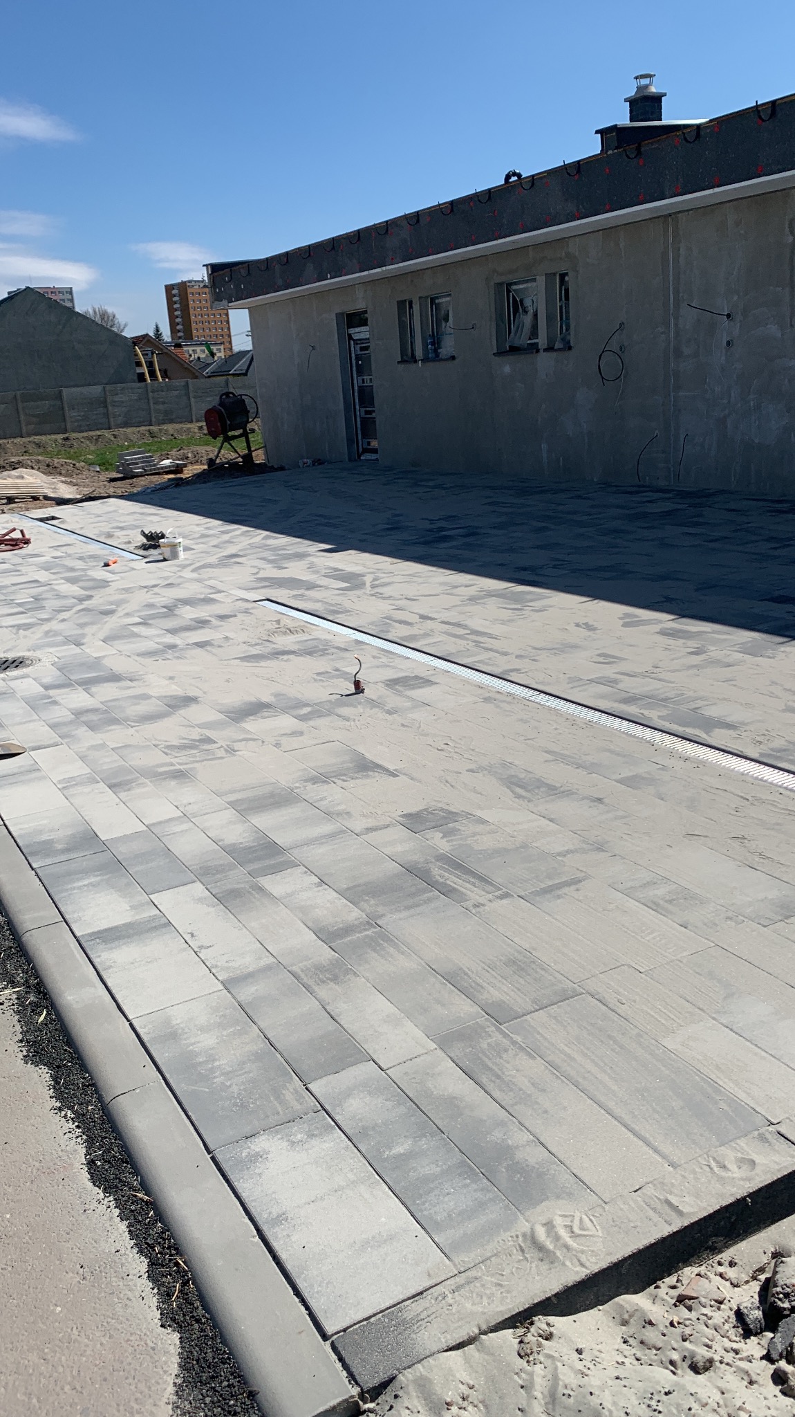 Ułożona szara kostka brukowa z odpływem liniowym na posesji. W tle budynek w stanie surowym i sprzęt budowlany. Słoneczny dzień.