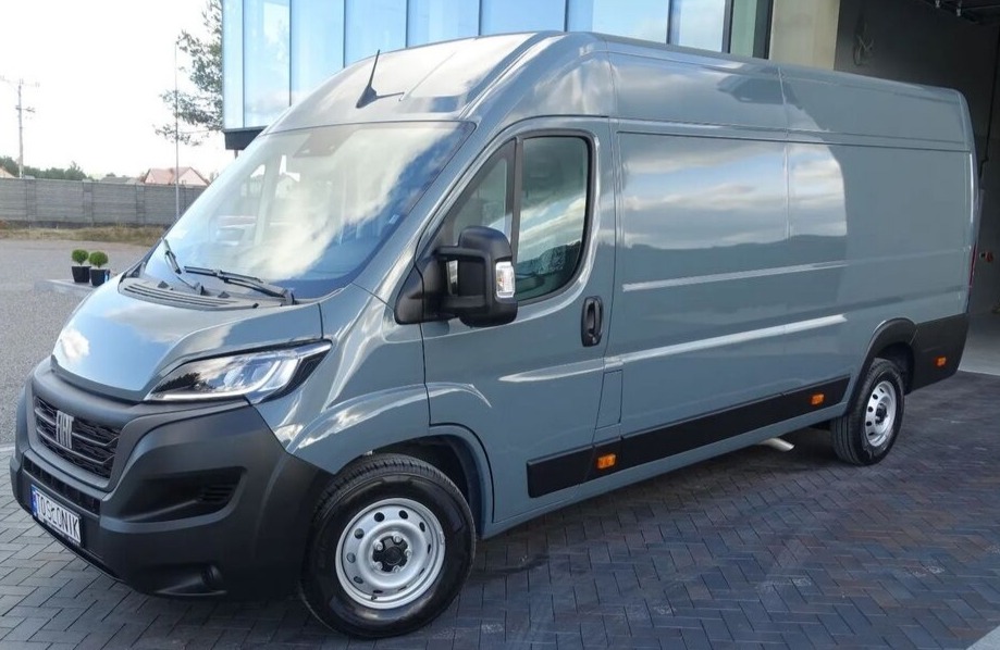 Szary samochód dostawczy Fiat Ducato zaparkowany na brukowanym podjeździe przed nowoczesnym budynkiem z szklaną fasadą.