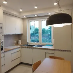 Jasna kuchnia z drewnianym stołem i krzesłami, widok na zieleń za oknem, nowoczesne oświetlenie sufitowe i wisząca lampa w stylu retro.