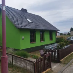 Świeżo pomalowany na jaskrawo zielono dom z ciemną dachówką, widoczne rusztowania za drewnianym płotem i skrzynką na listy, pochmurne niebo.