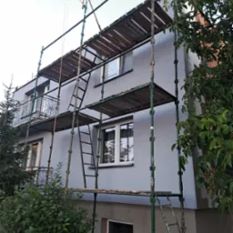 Ocieplony szary budynek dwupiętrowy w trakcie malowania, otoczony zielonym rusztowaniem z drewnianymi pomostami i małą wciągarką linową na dachu.