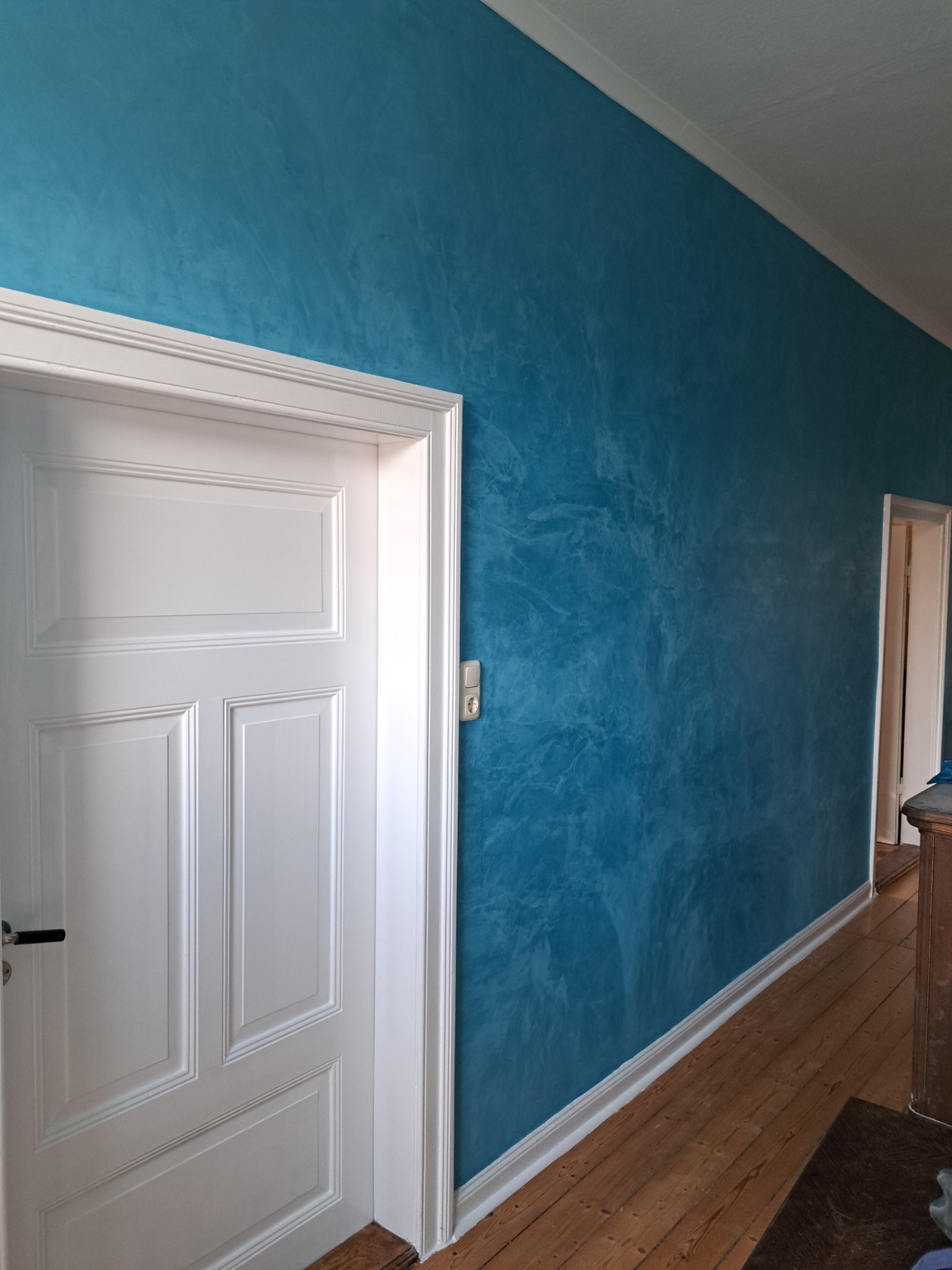 Błękitna ściana z efektem dekoracyjnym, widoczna obok białych drzwi z panelami i drewnianej podłogi, w tle fragment korytarza.