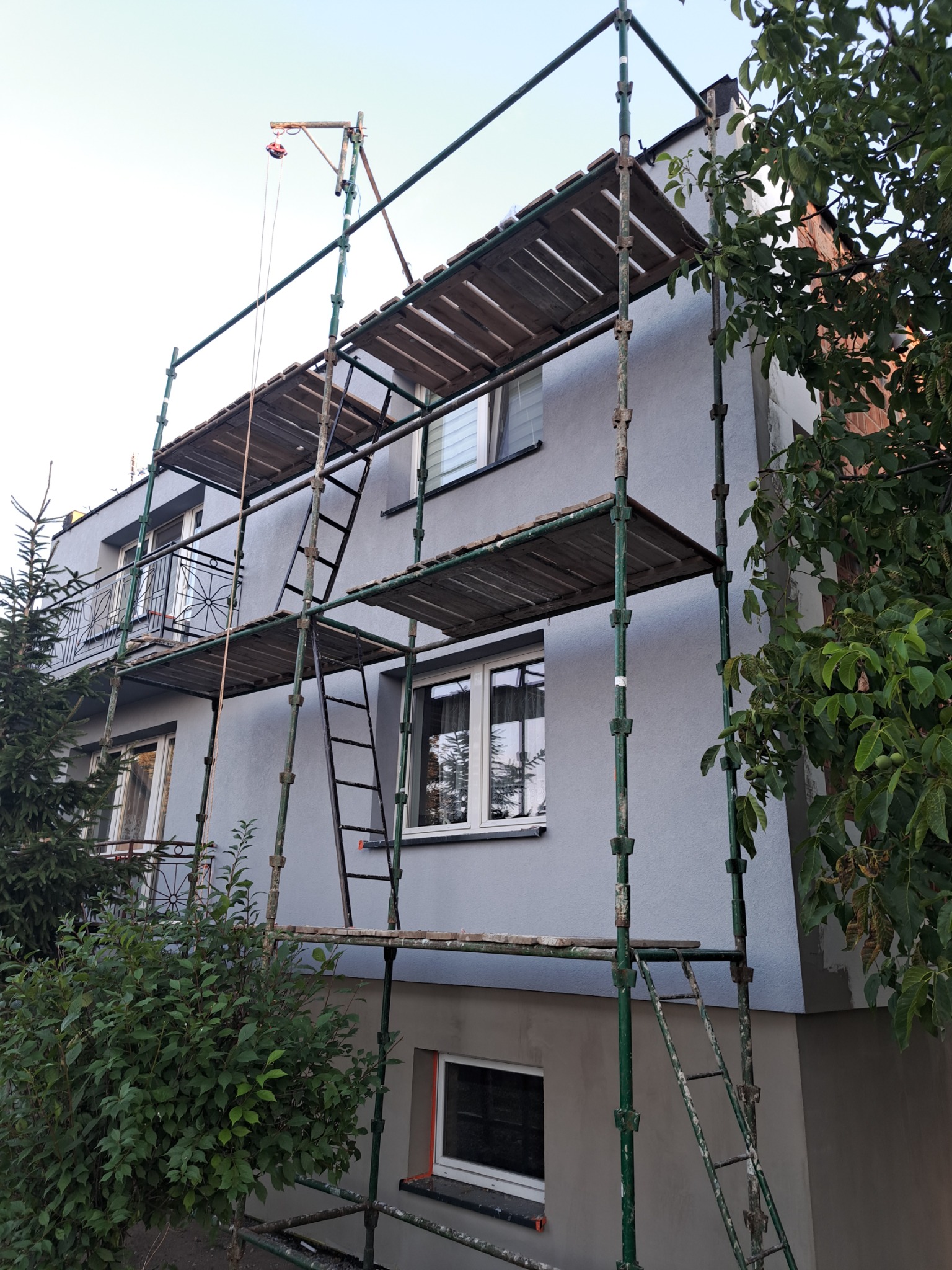 Ocieplony szary budynek dwupiętrowy w trakcie malowania, otoczony zielonym rusztowaniem z drewnianymi pomostami i małą wciągarką linową na dachu.