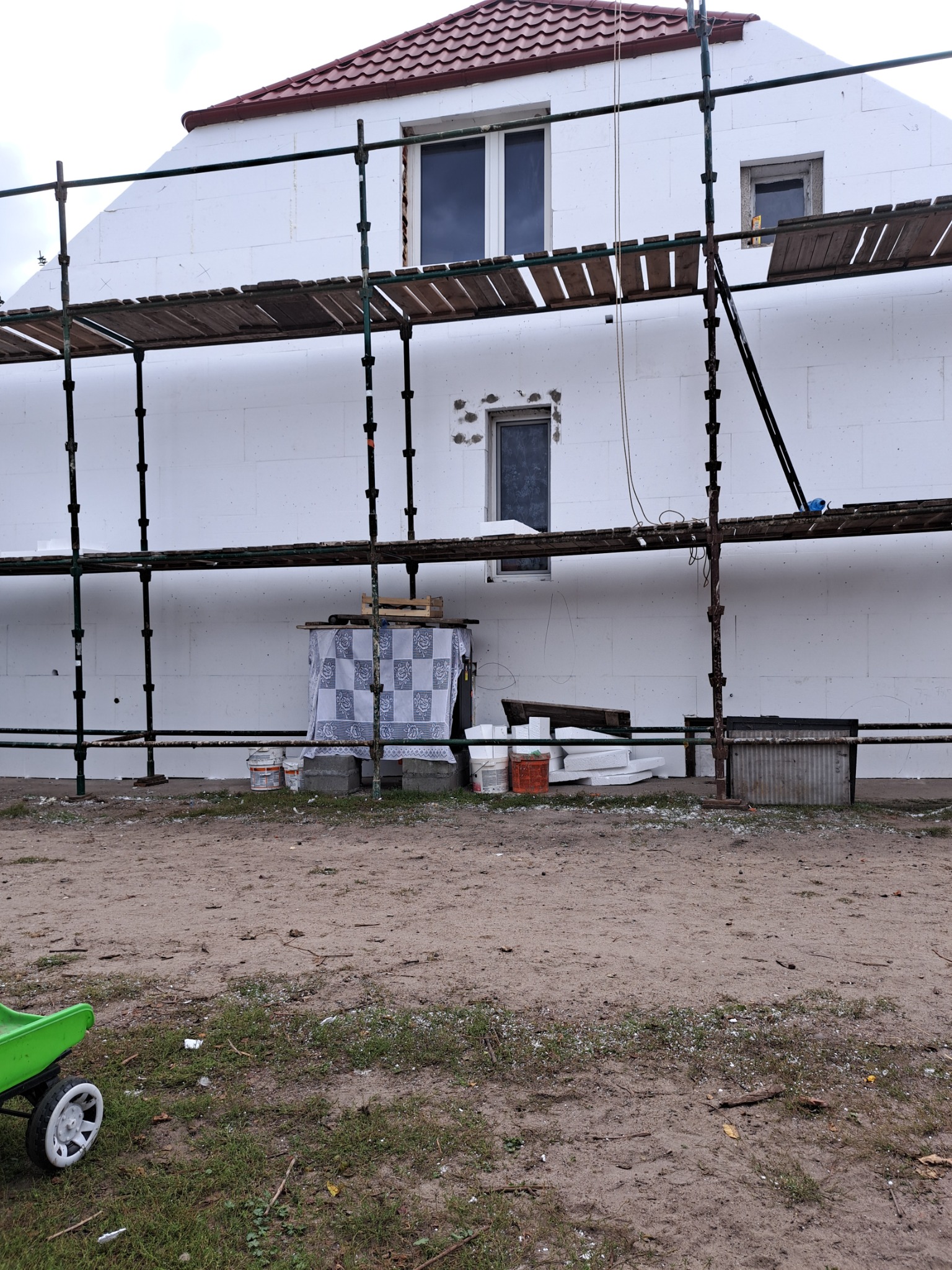 Dom w trakcie ocieplania styropianem, widoczne rusztowania, okna, częściowo ocieplona ściana i teren budowy z zielonym wózkiem.