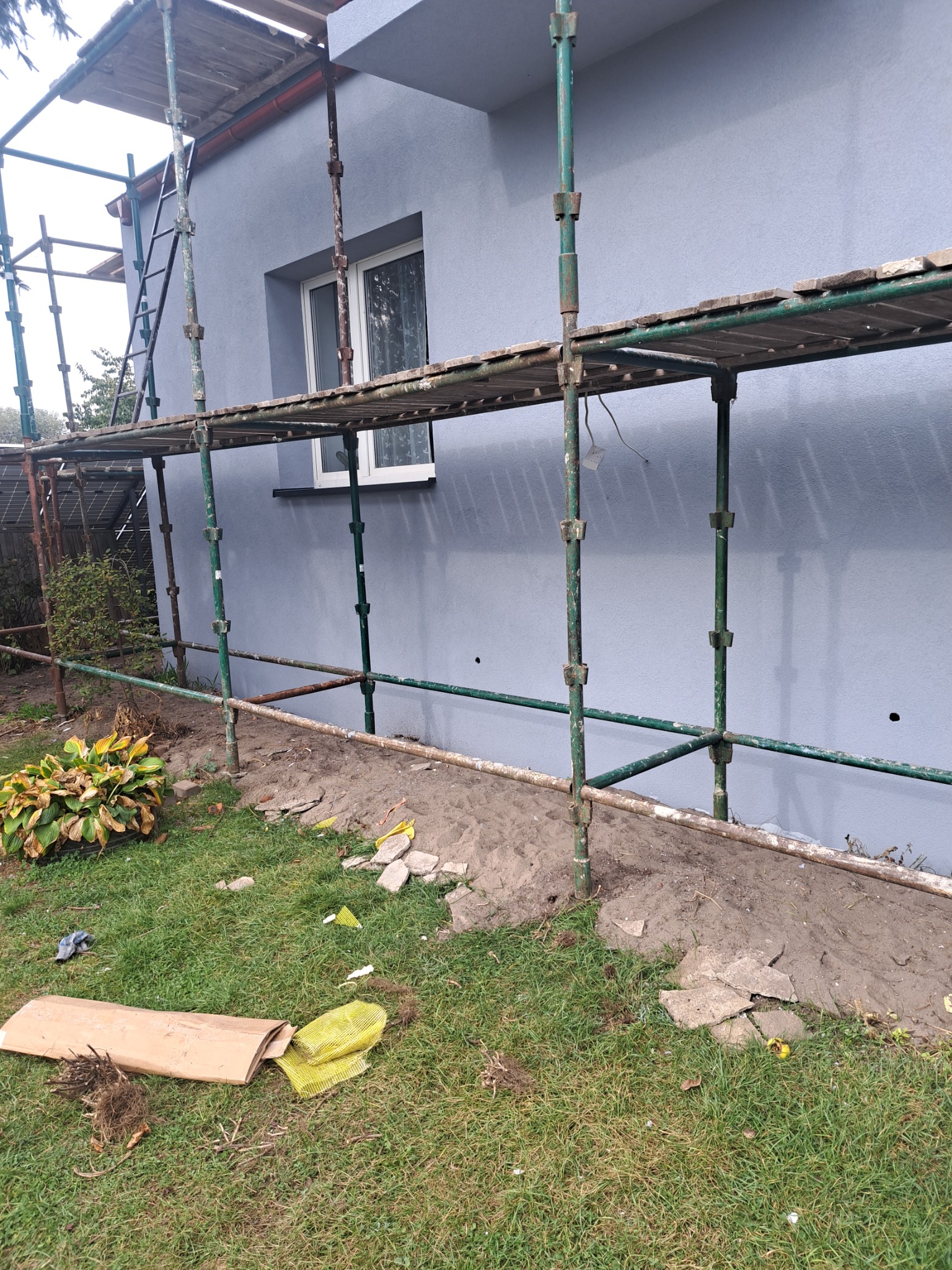 Szare wykończenie elewacji domu jednorodzinnego, widoczne z poziomu gruntu z zielonymi rusztowaniami i drabiną, fragment okna i roślinności w tle, oraz resztki gruzu i odpadów budowlanych na trawie.