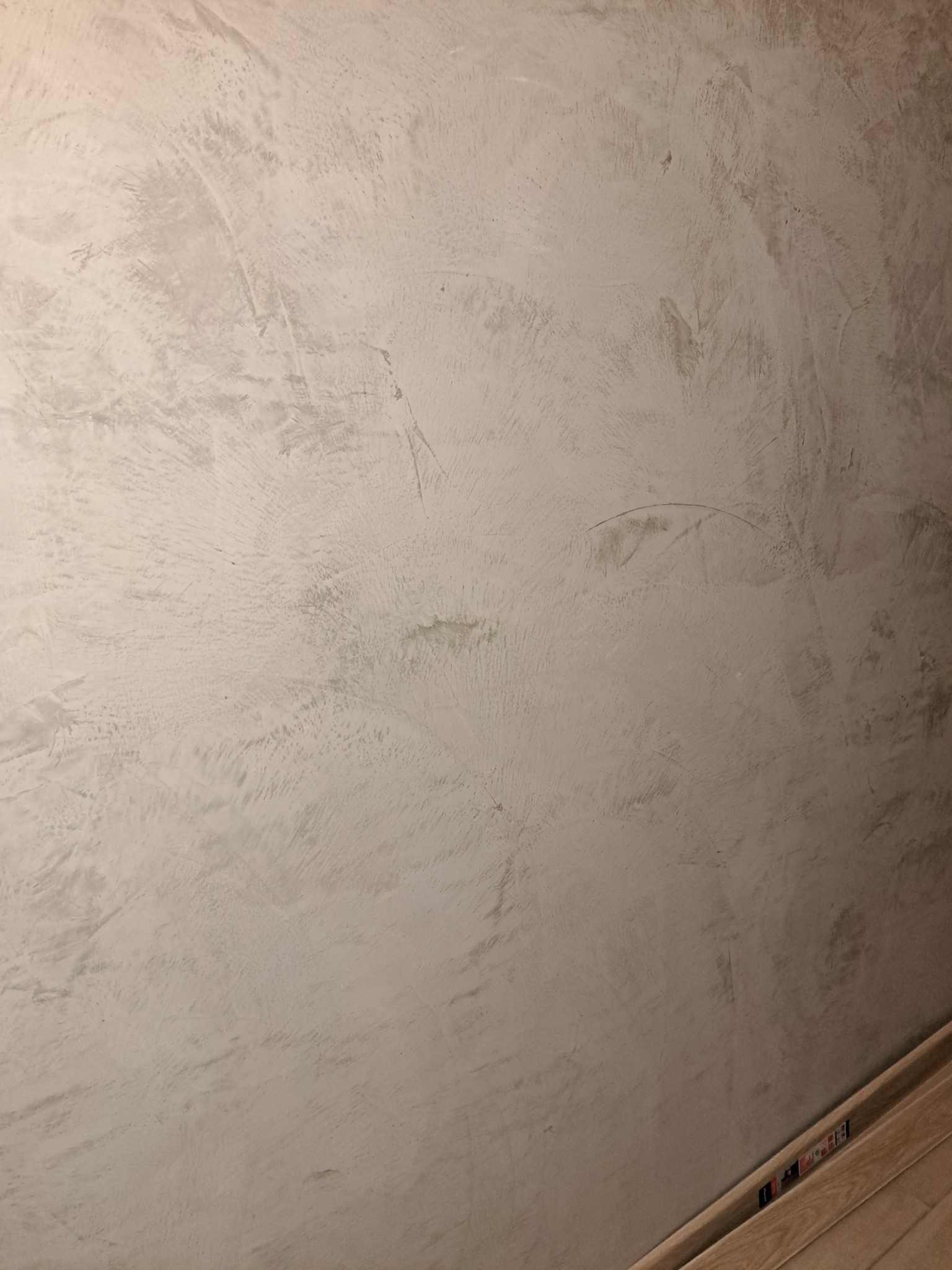 Ściana pokryta szarą, dekoracyjną strukturą z widocznymi śladami narzędzi, oświetlona z góry, z widoczną przypodłogową listwą wykończeniową.