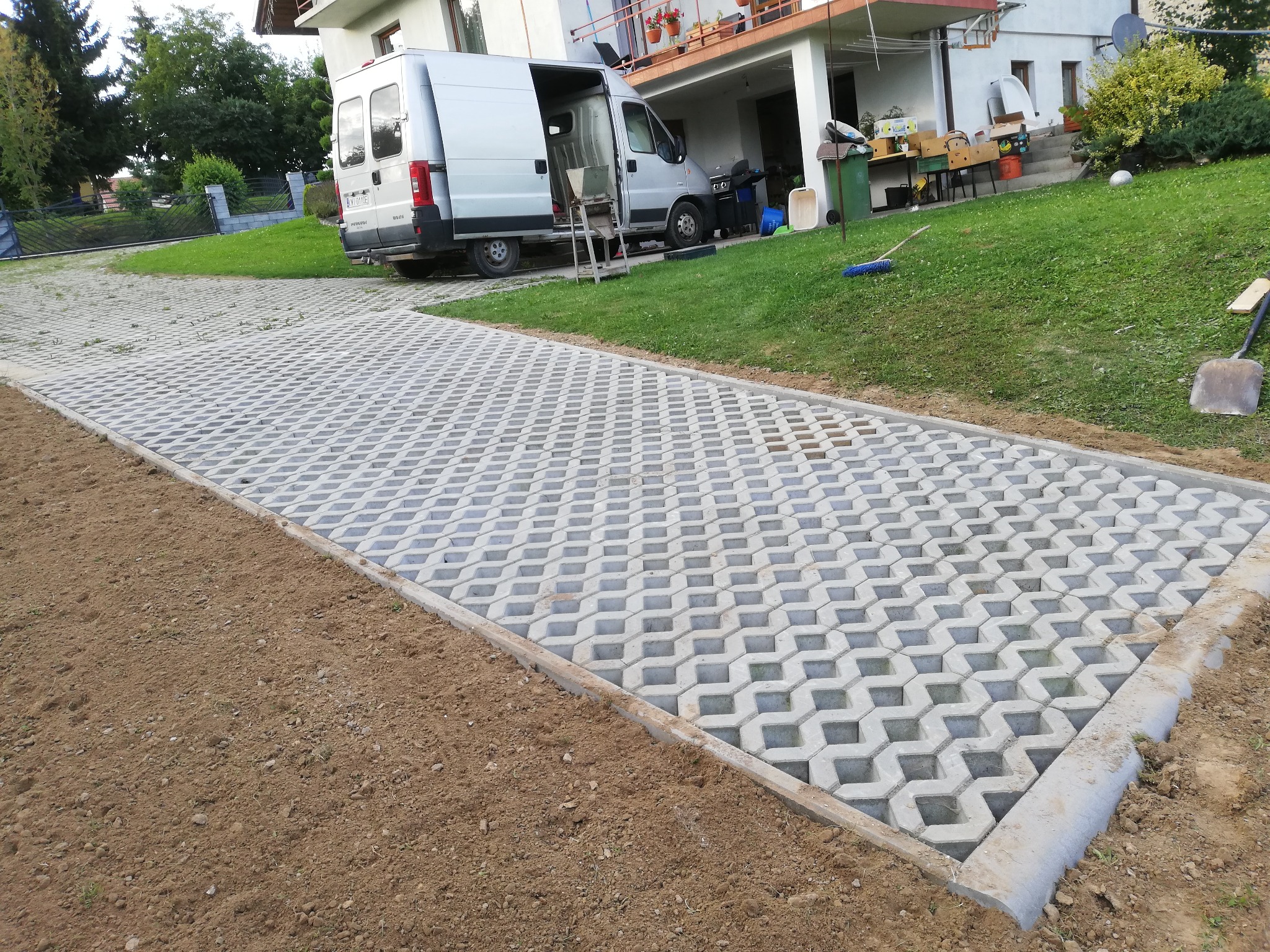 Układanie przepuszczalnej kostki brukowej na podjeździe, widoczny srebrny bus z otwartymi drzwiami, ziemia przygotowana pod dalsze prace.