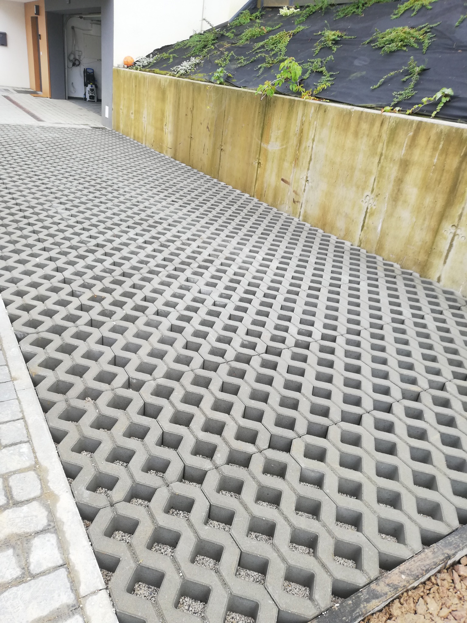 Szara ażurowa nawierzchnia z kostki betonowej wypełniona drobnym żwirem, prowadząca w kierunku wjazdu do garażu, z widocznym murem oporowym obsadzonym roślinnością w tle.