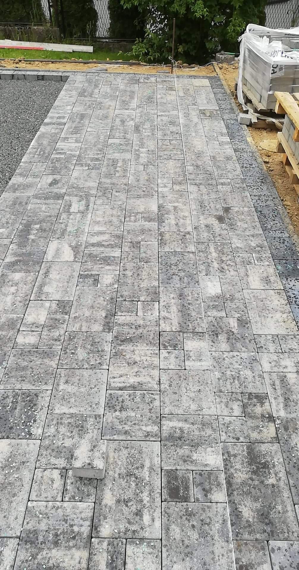 Ułożona ścieżka z szarej kostki brukowej o nieregularnym wzorze, z widocznymi zapasami kostki na palecie w tle i przygotowanym podłożem z boku.