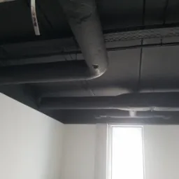 Pomieszczenie biurowe z pomalowanym na czarno sufitem, na którym widoczne są rury i instalacje; białe ściany i okno w tle.