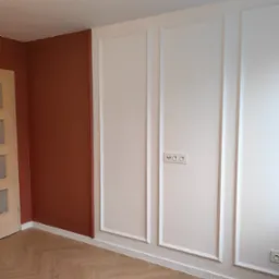 Pokój po remoncie: ściana w kolorze terakoty, białe panele ścienne z dekoracyjnymi listwami, parkiet ułożony w jodełkę, jasne drzwi.