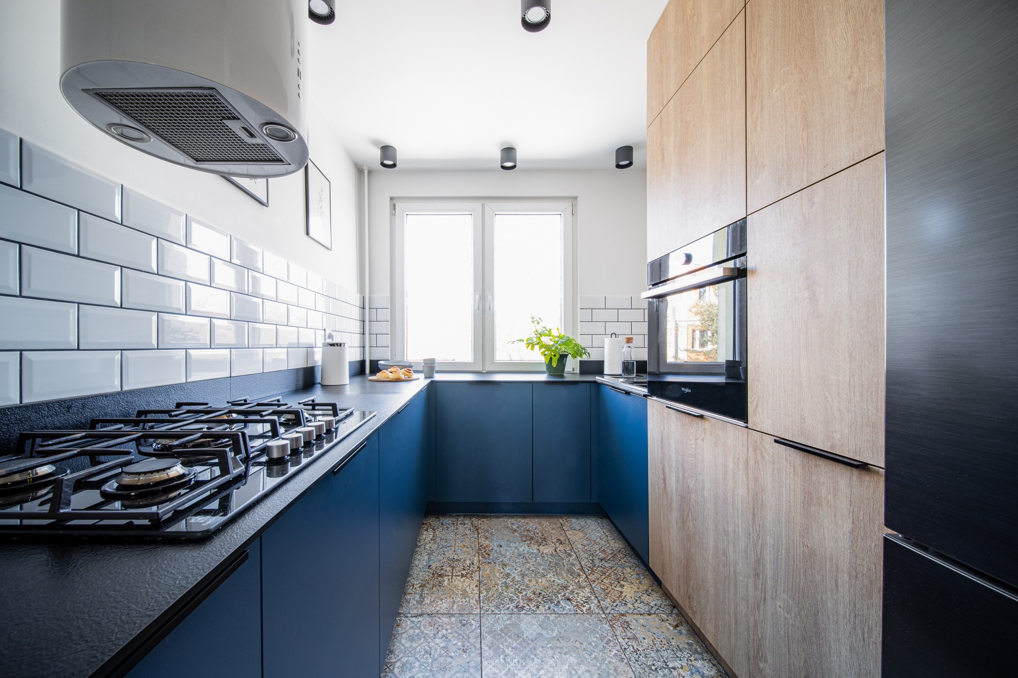 Nowoczesna kuchnia w kształcie litery U z niebieskimi szafkami, drewnopodobnymi frontami, czarnym blatem i białą cegłą na ścianie, oświetlona punktowymi lampami sufitowymi i naturalnym światłem...