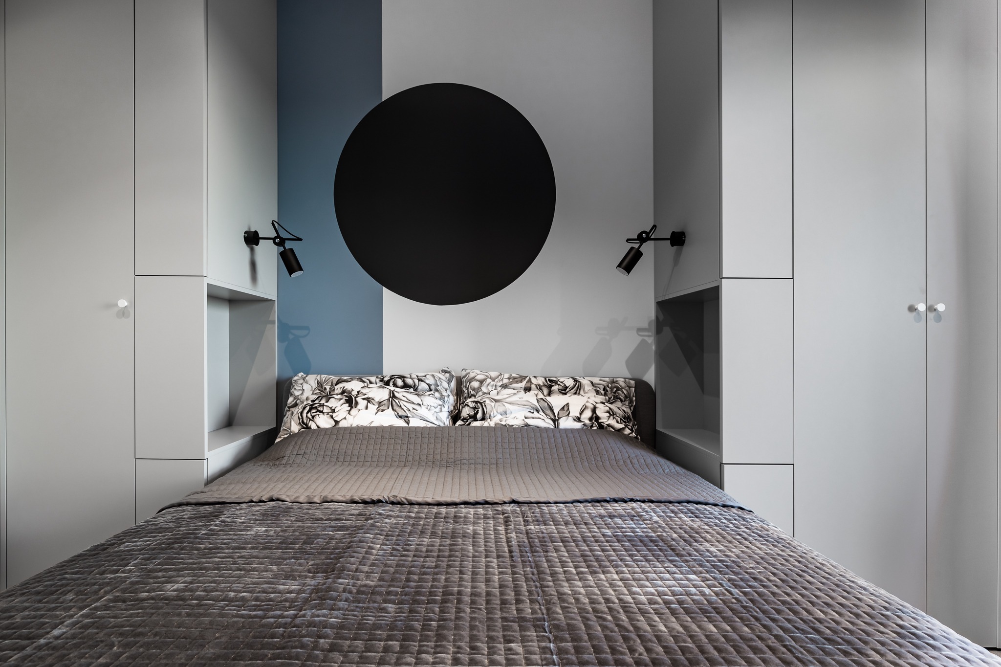 Nowoczesna sypialnia z szarą pościelą w kwadraty, poduszkami w kwiaty, czarnym okręgiem na ścianie i minimalistycznymi lampkami nocnymi, otoczona zabudową meblową w odcieniach szarości.