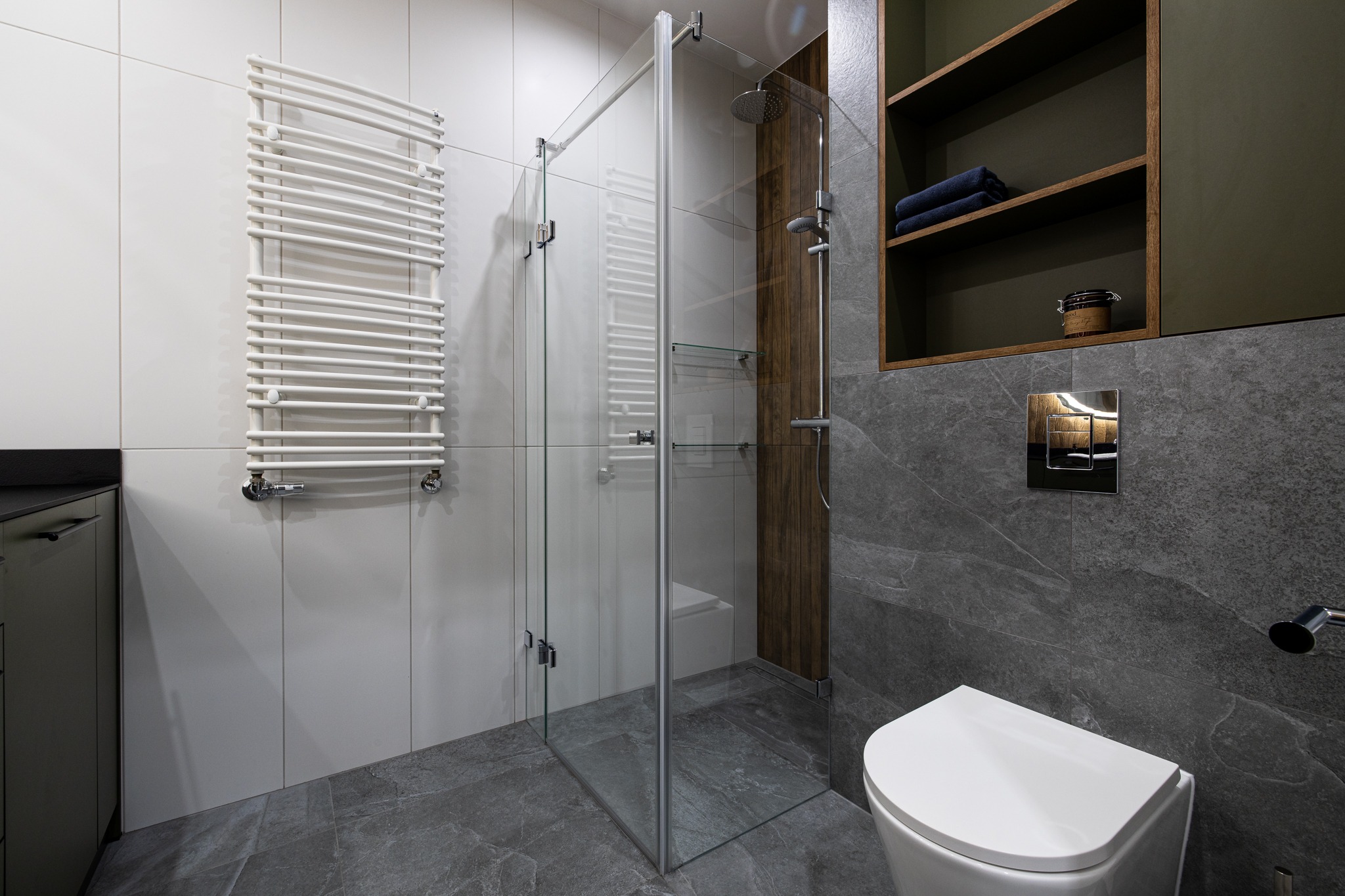 Nowoczesna łazienka z kabiną prysznicową ze szklanymi ścianami, grzejnikiem drabinkowym na białej ścianie z płytkami, szarymi płytkami imitującymi kamień na podłodze i ścianie za toaletą...