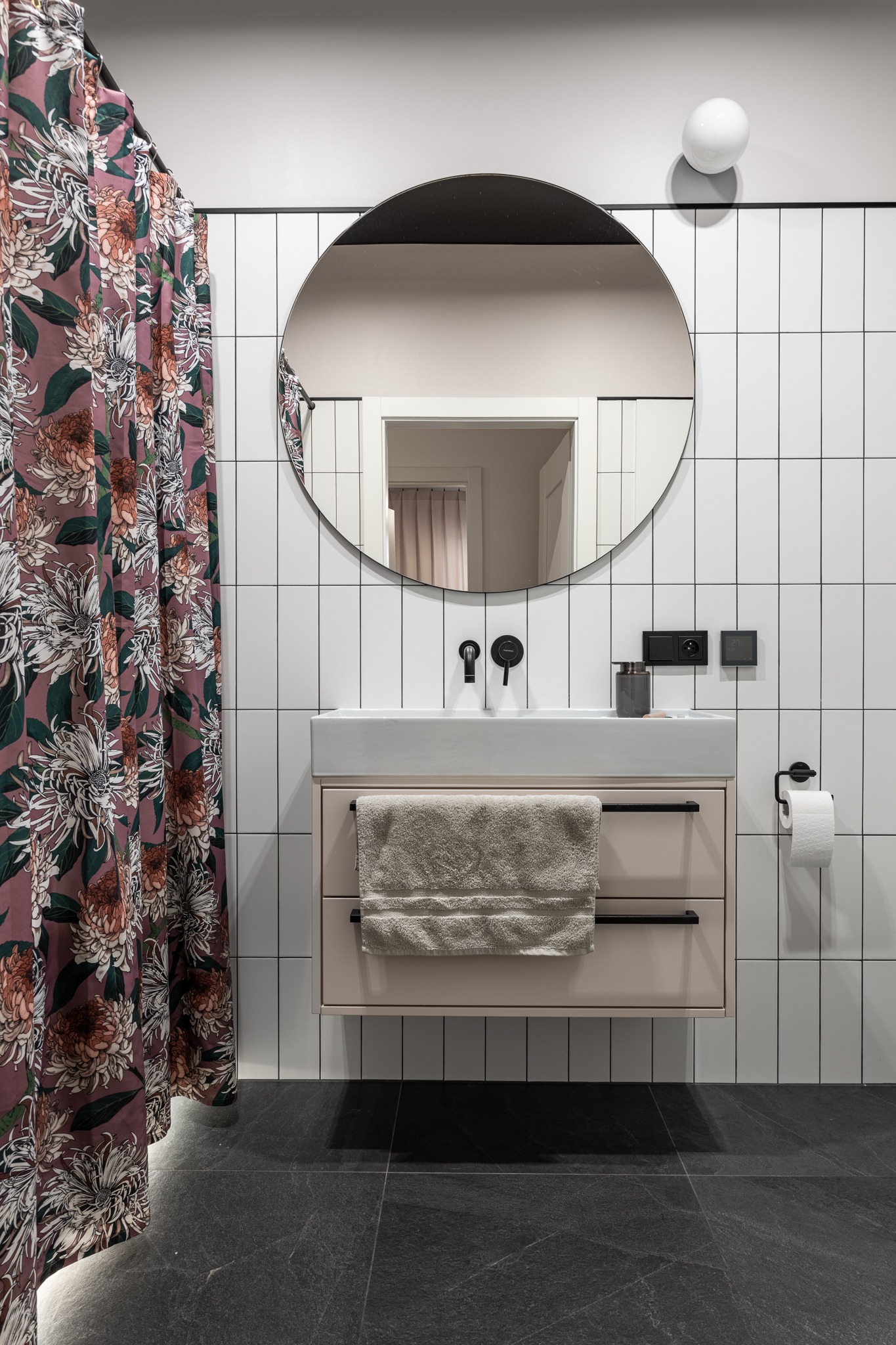 Nowoczesna łazienka z wiszącą szafką umywalkową w kolorze pudrowego różu, okrągłym lustrem z czarną ramą, czarną armaturą i białą pionową glazurą, uzupełniona zasłoną prysznicową w kwiatowy wzór.