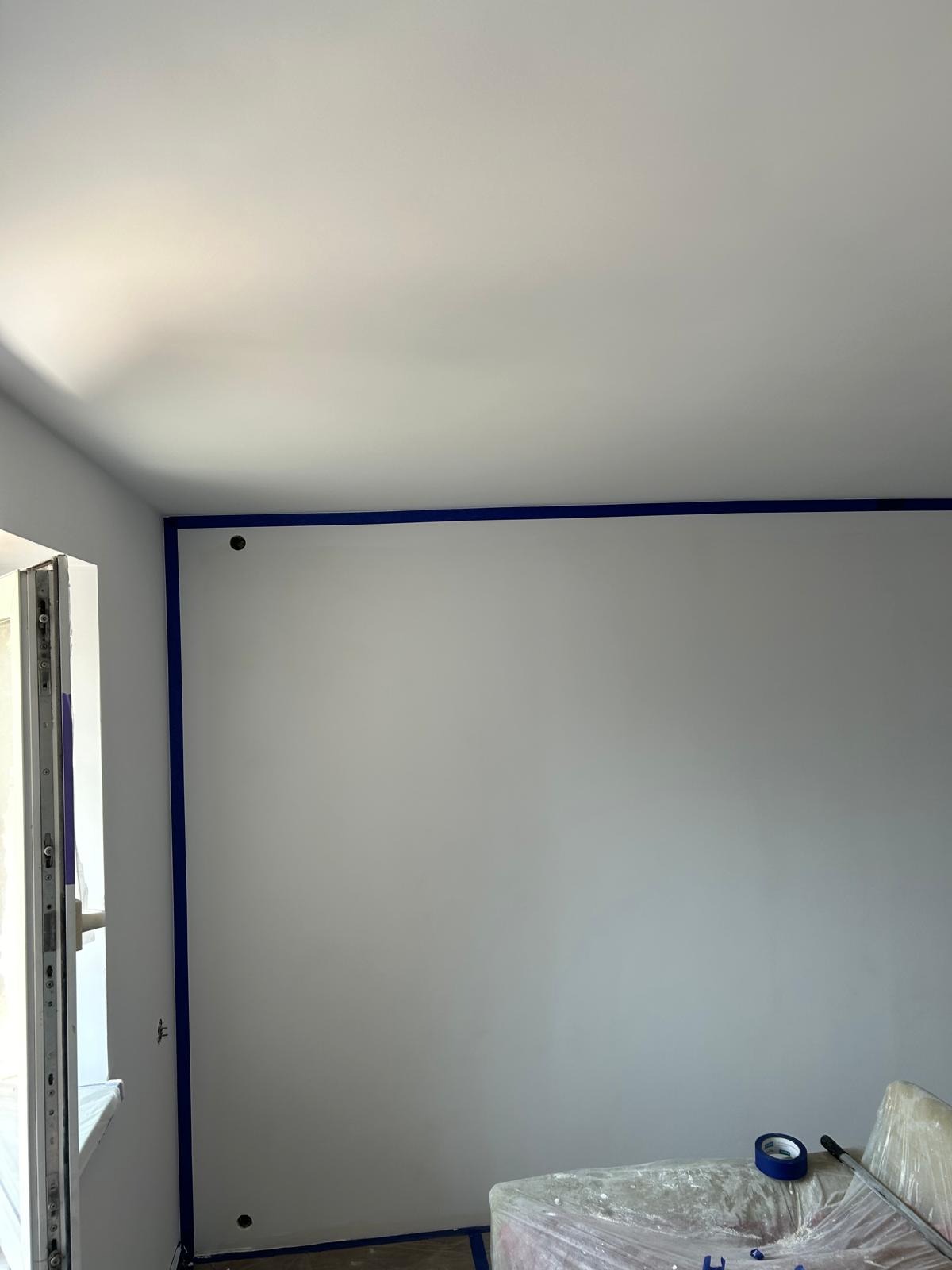 Przygotowanie do malowania: świeżo pomalowane ściany i sufit na biało, zabezpieczone taśmą malarską wzdłuż krawędzi i przy podłodze, widoczne puszki po farbie i folia ochronna na podłodze.