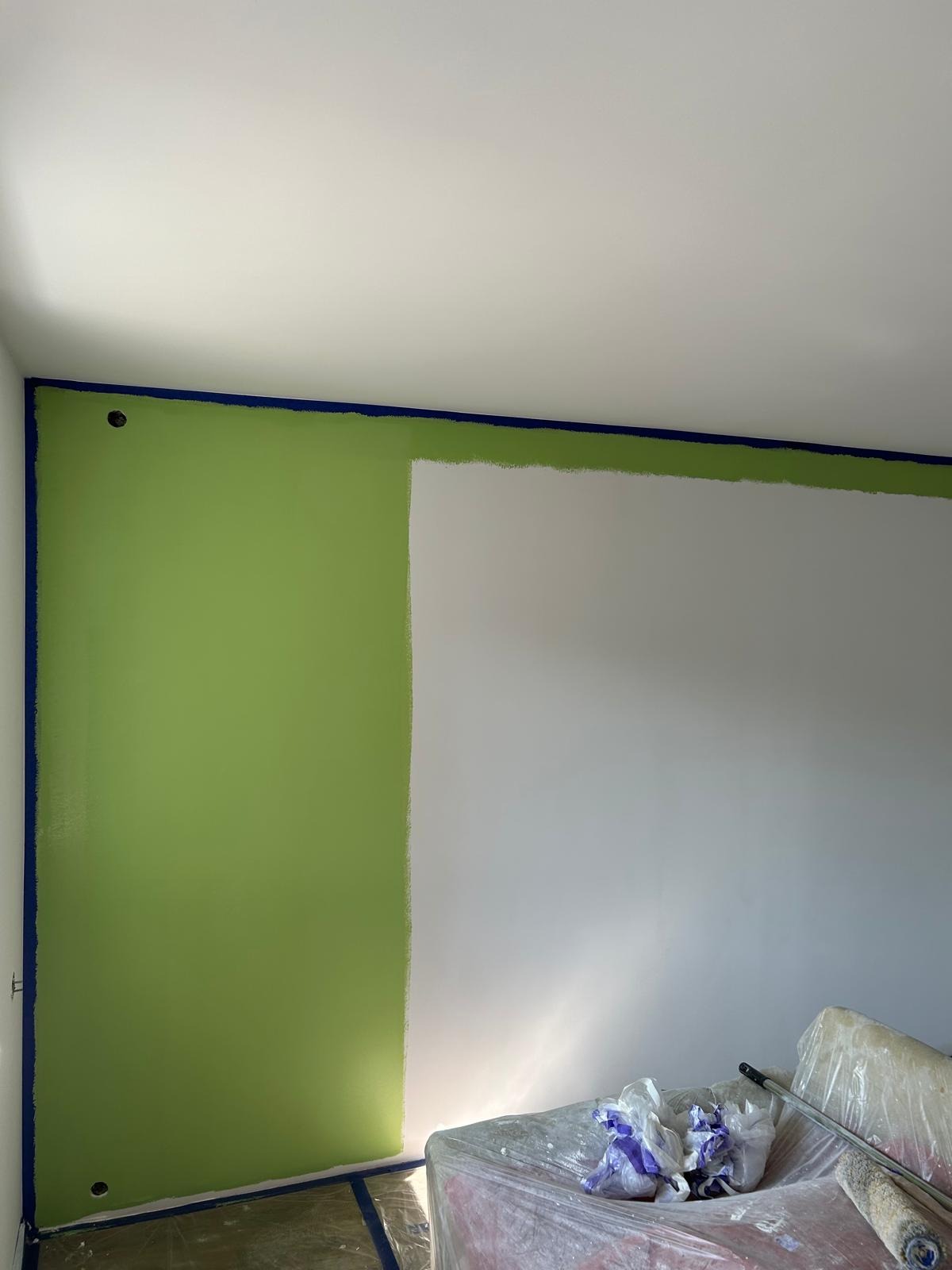 Wnętrze pokoju w trakcie malowania: ściana częściowo pomalowana na jasnozielony kolor, druga część na biało, zabezpieczona taśmą malarską, z widocznymi narzędziami malarskimi przykrytymi folią.