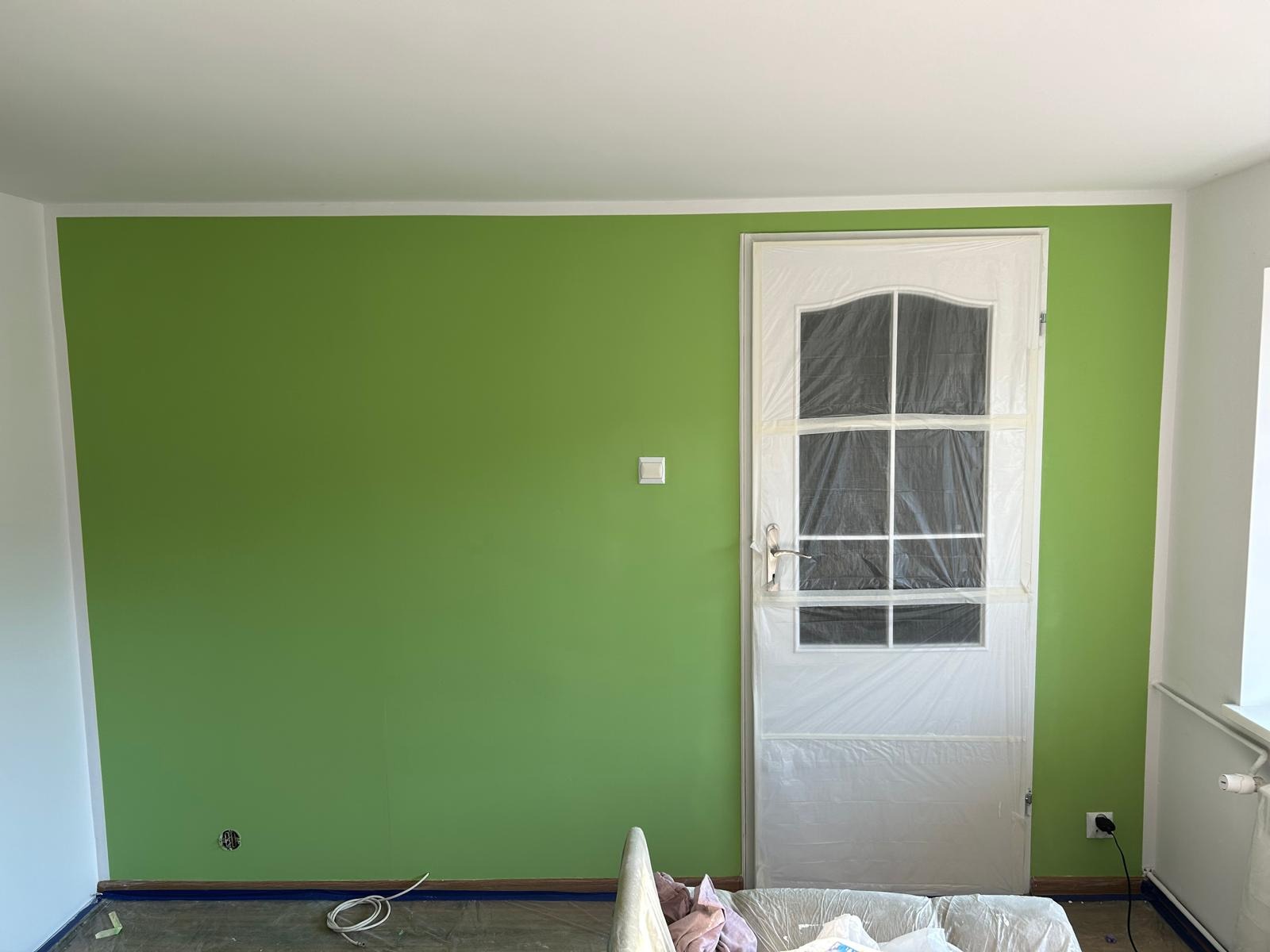 Świeżo pomalowana ściana na zielono w pokoju, zabezpieczone drzwi folią malarską, widoczne ślady prac malarskich na podłodze.