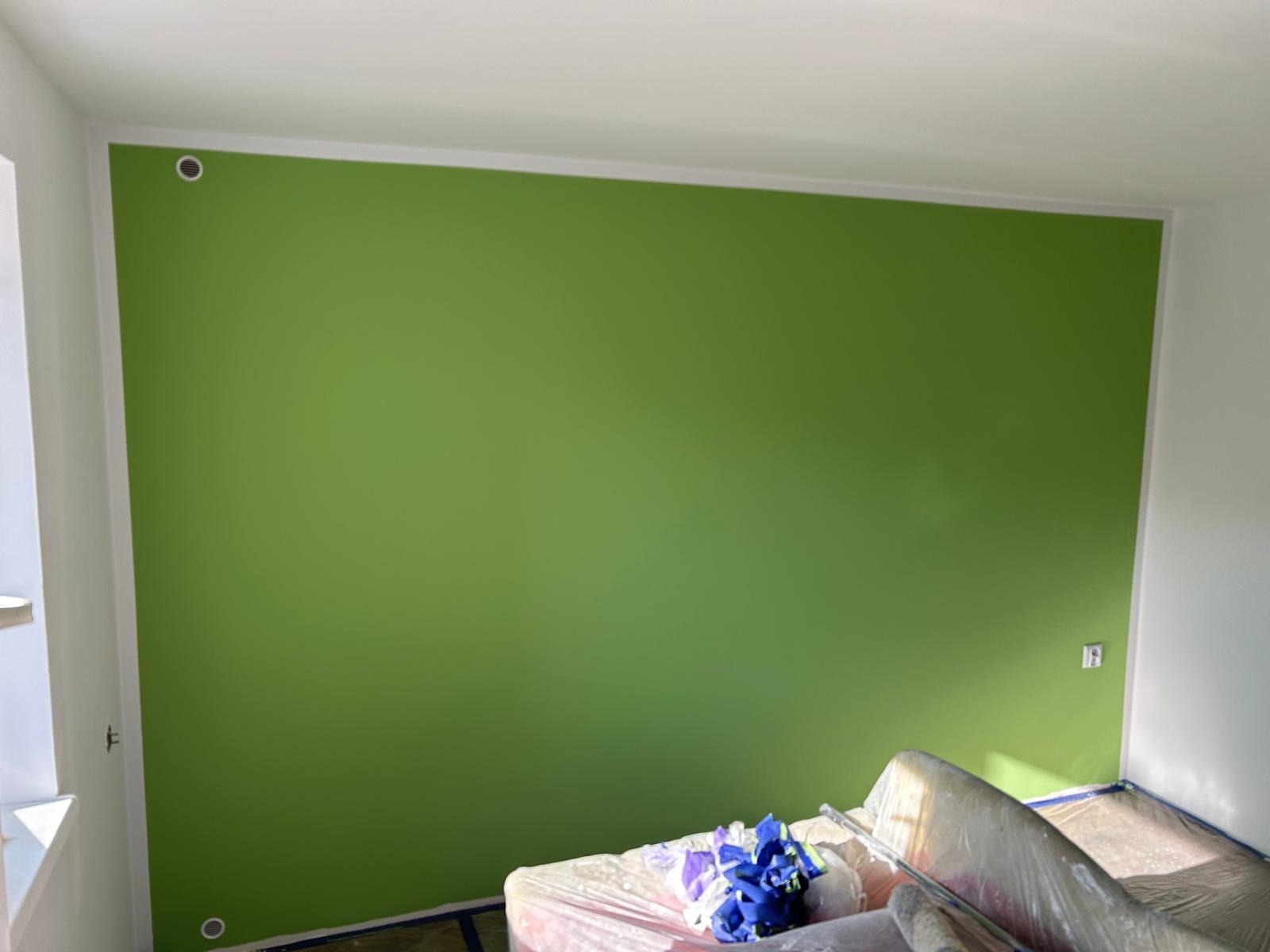 Świeżo pomalowana na zielono ściana w pokoju z widocznymi zabezpieczeniami podłogi i mebli folią malarską, dwa otwory wentylacyjne w górnej i dolnej części ściany.