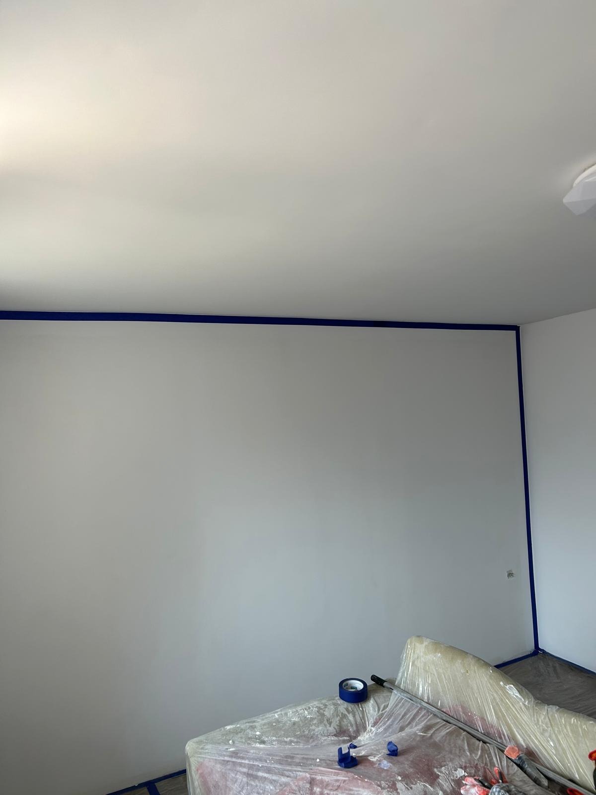 Przygotowanie do malowania: Świeżo pomalowana, jasnoszara ściana z niebieską taśmą malarską przy suficie i narożnikach, zabezpieczone meble folią malarską, widoczna rolka i taśma malarska.